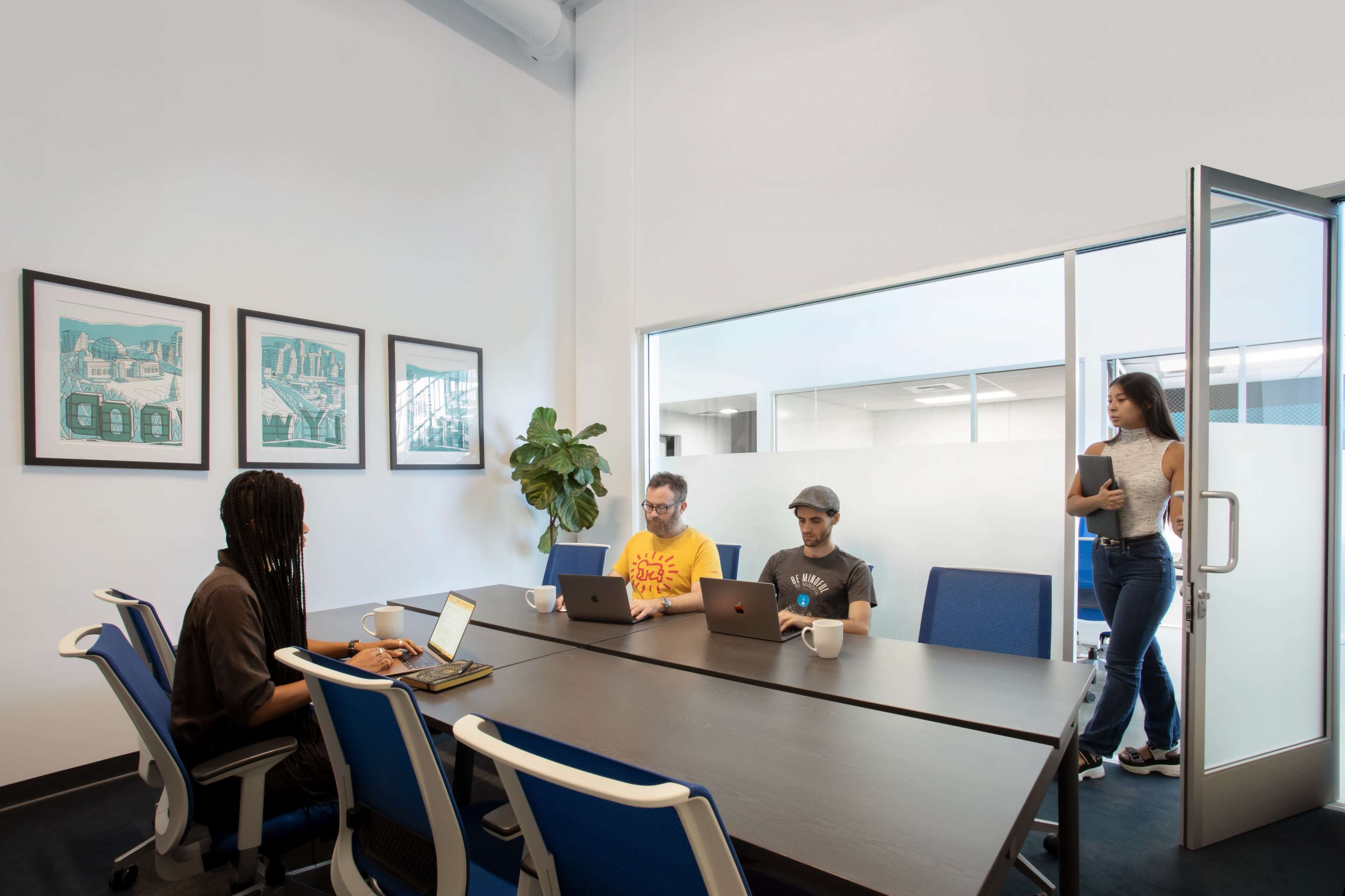A group of four people works in a modern conference room, with two individuals at a table using laptops and a woman entering through a glass door.