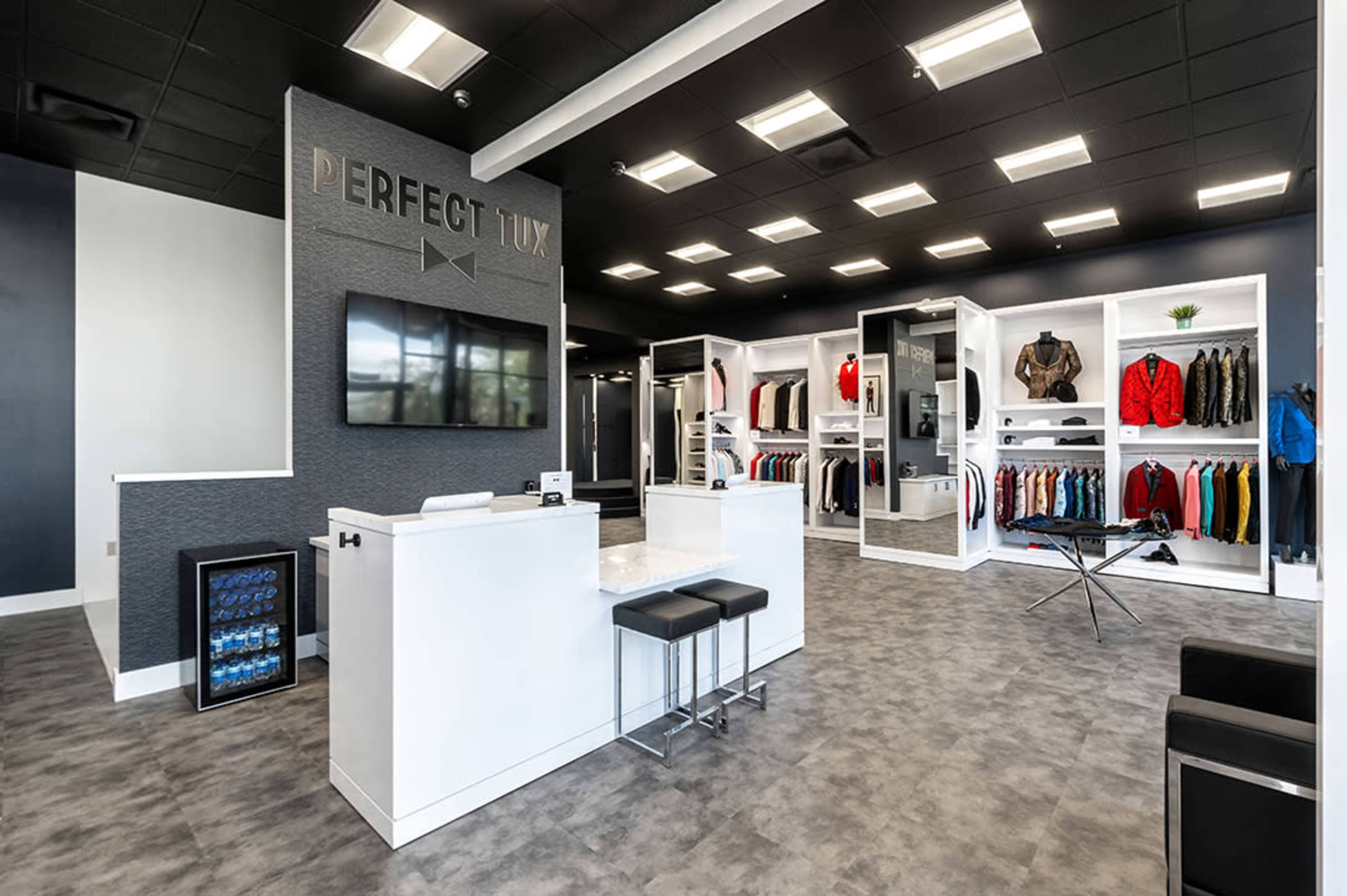 The interior of a tuxedo rental shop featuring a reception area with a white counter and seating, alongside neatly arranged clothing displays.