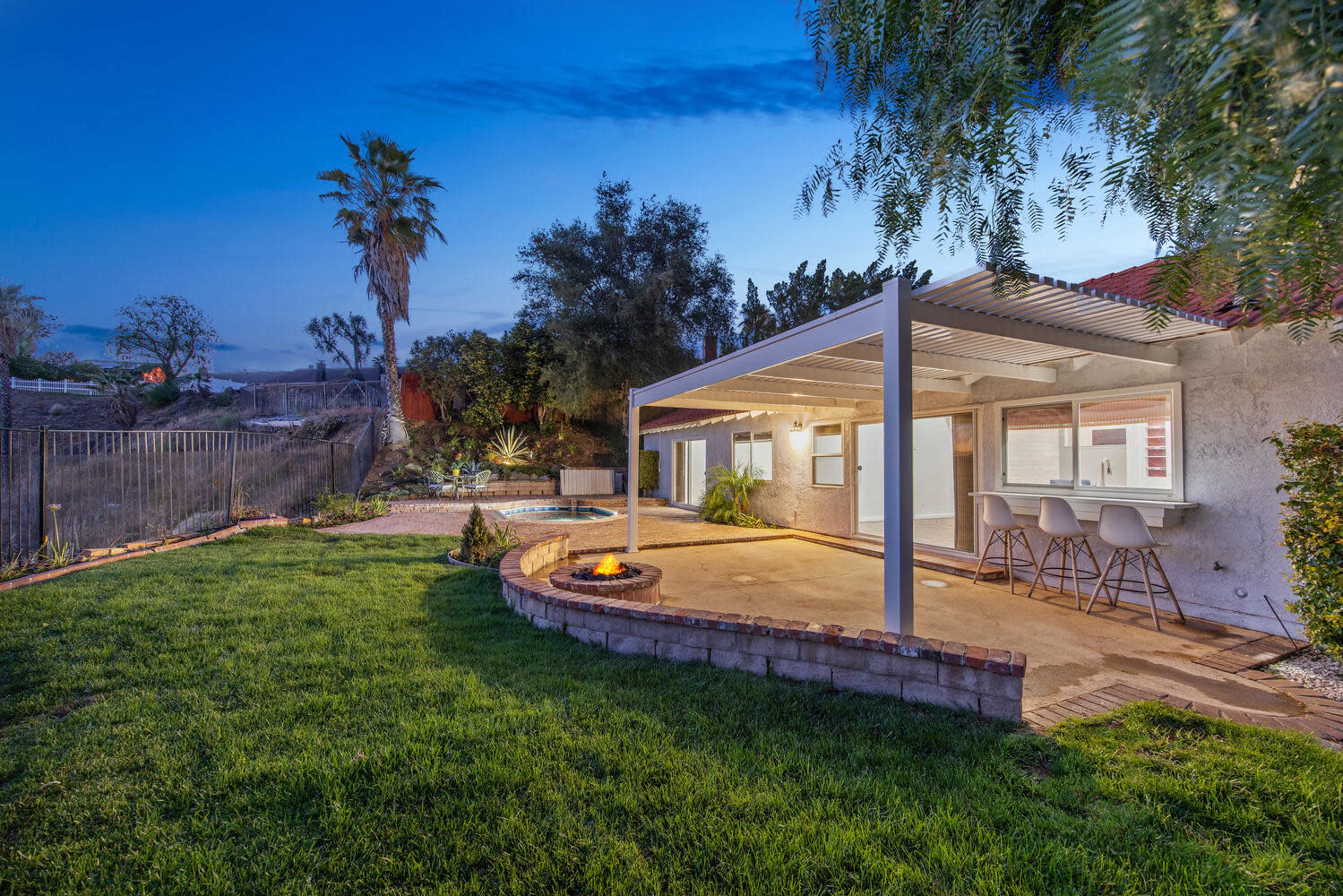 A backyard features a covered patio with seating, a fire pit, and a pool surrounded by greenery and palm trees at dusk.