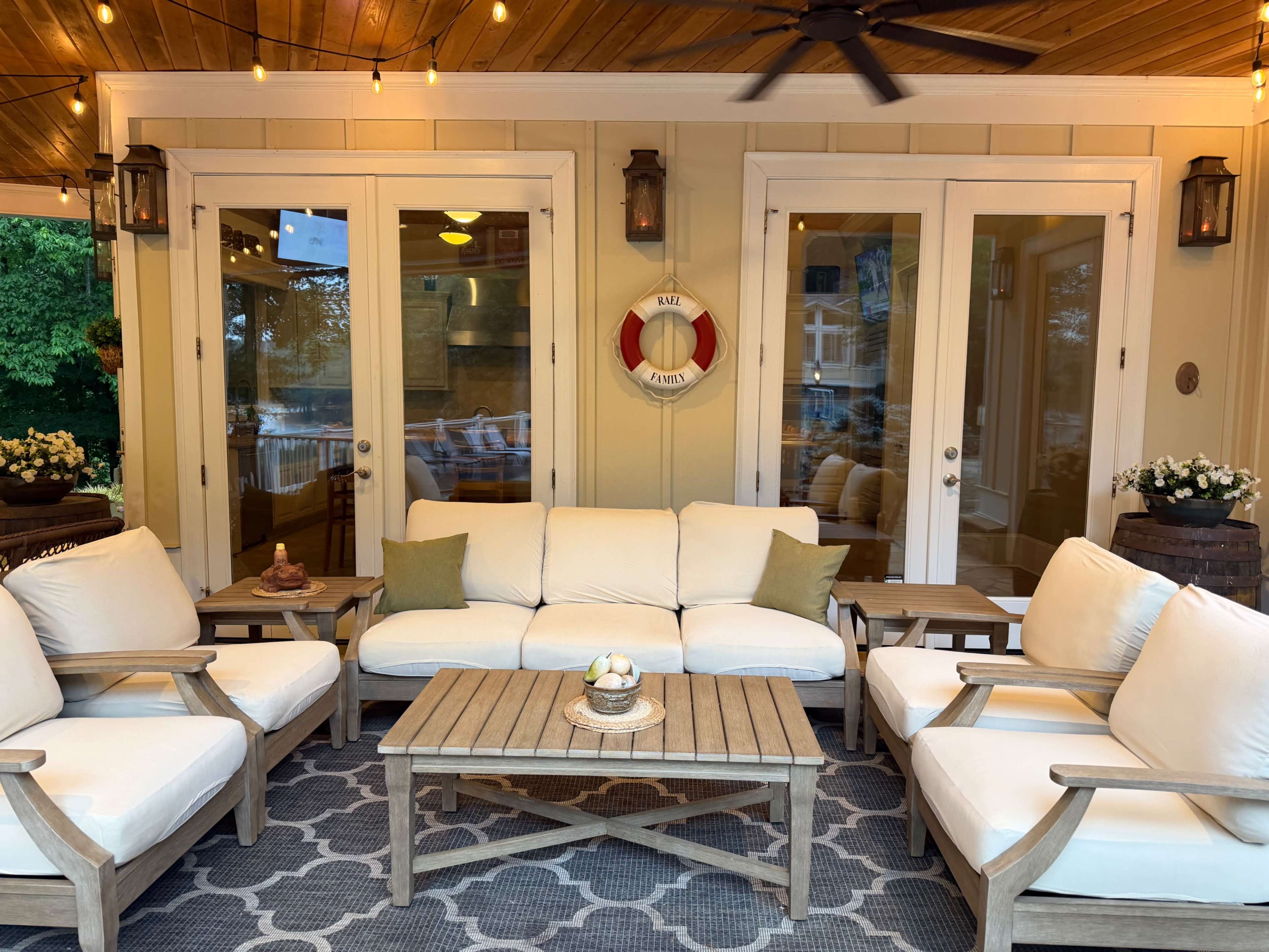 The image shows a cozy outdoor seating area with white cushions on wooden chairs, a wooden table in the center, and decorative lighting hanging above.