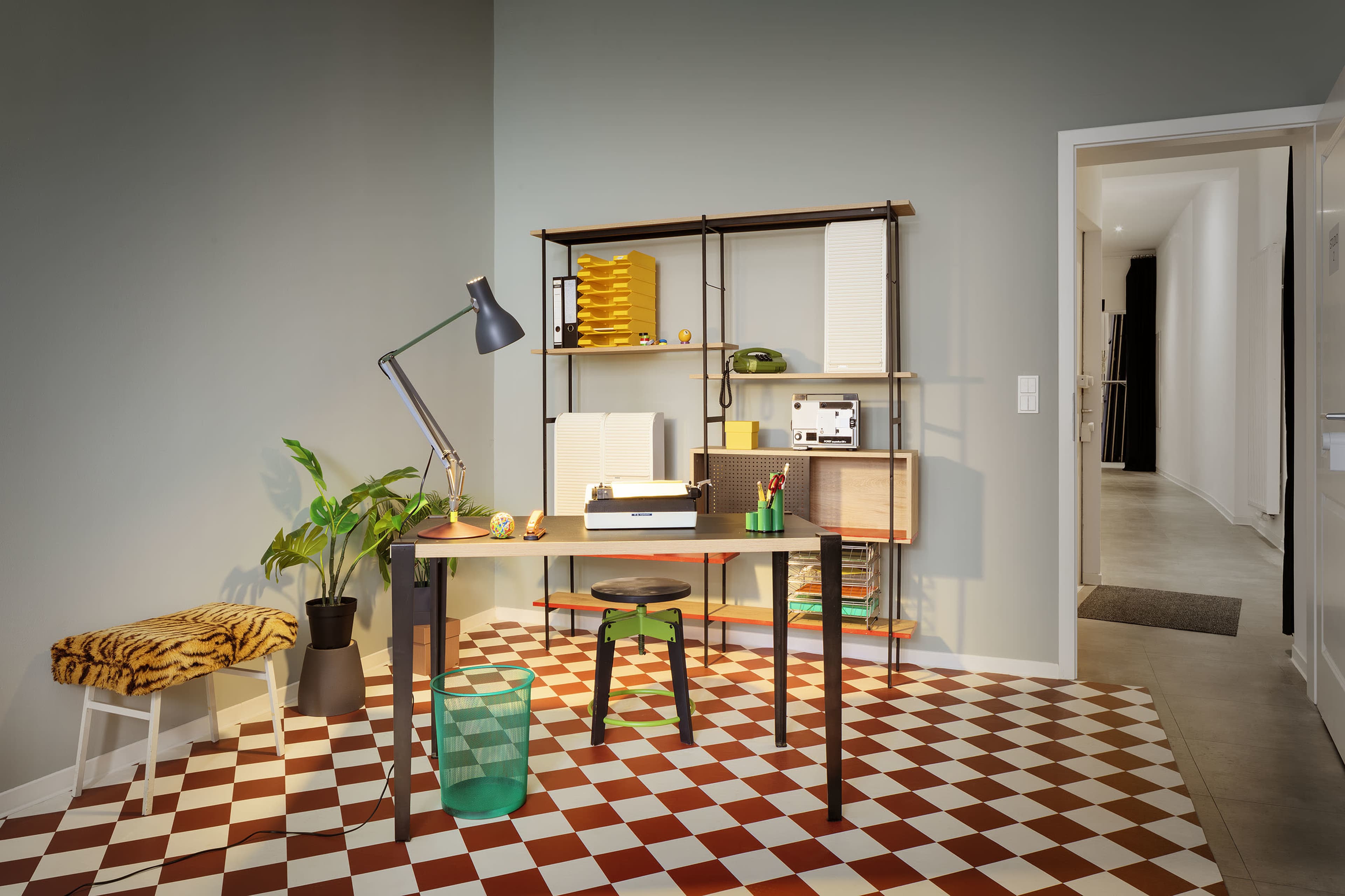 A modern workspace features a desk with a lamp, a plant, and a shelving unit against a green wall, with a tiled floor in red and white.