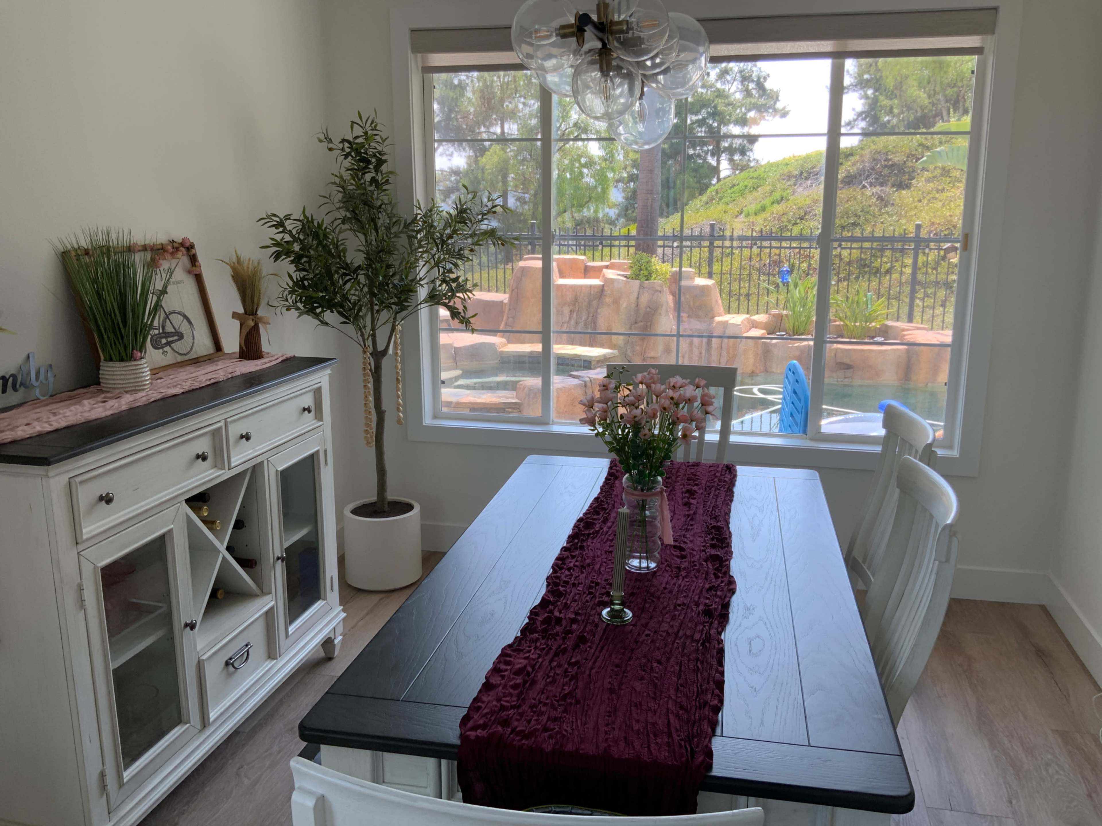 A dining area features a wooden table with a burgundy table runner, surrounded by white chairs, and looks out over a pool through a large window.