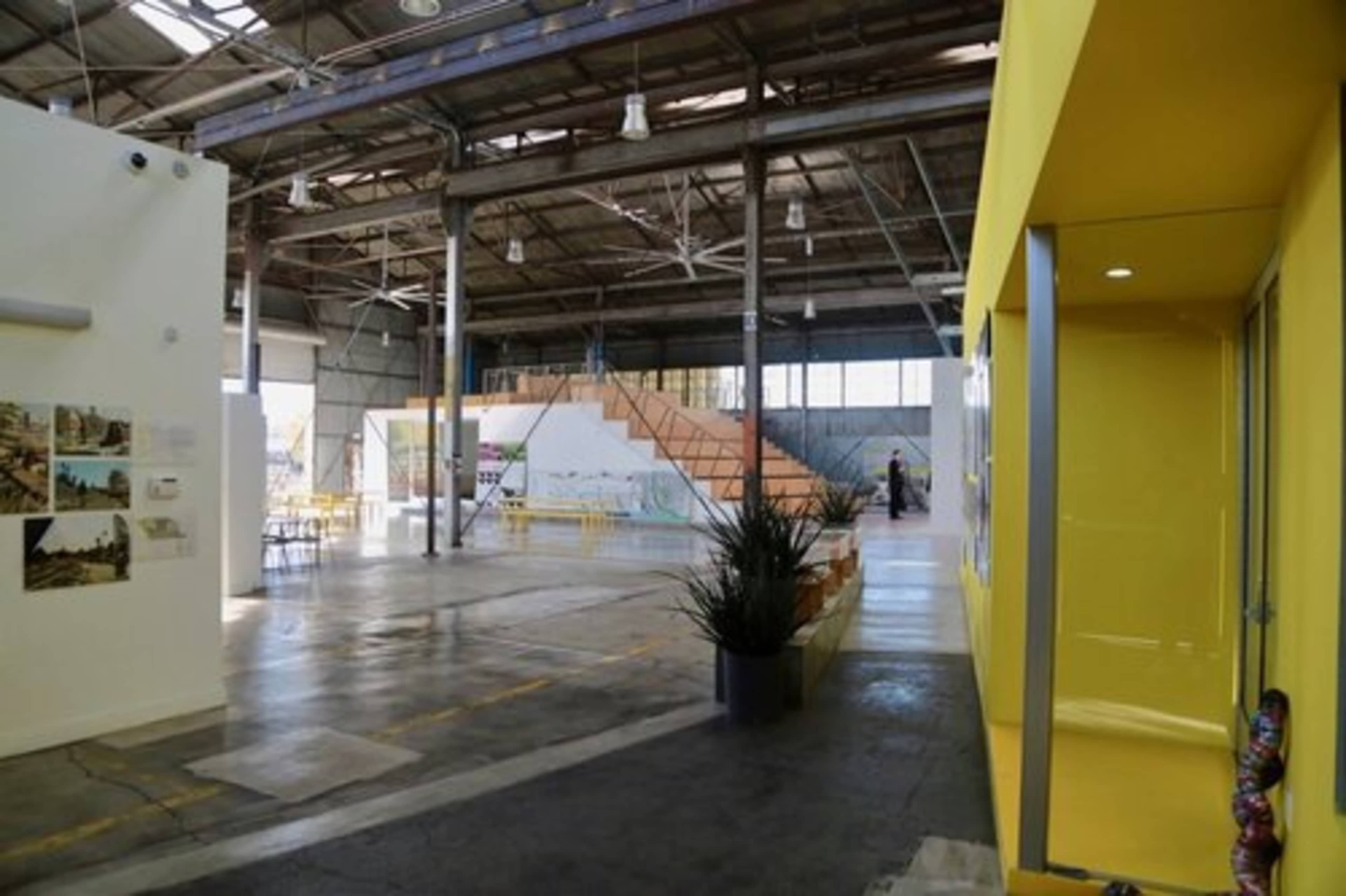 The image shows an industrial building interior with high ceilings, featuring a yellow entrance on the right and a staircase in the distance leading to an elevated area.