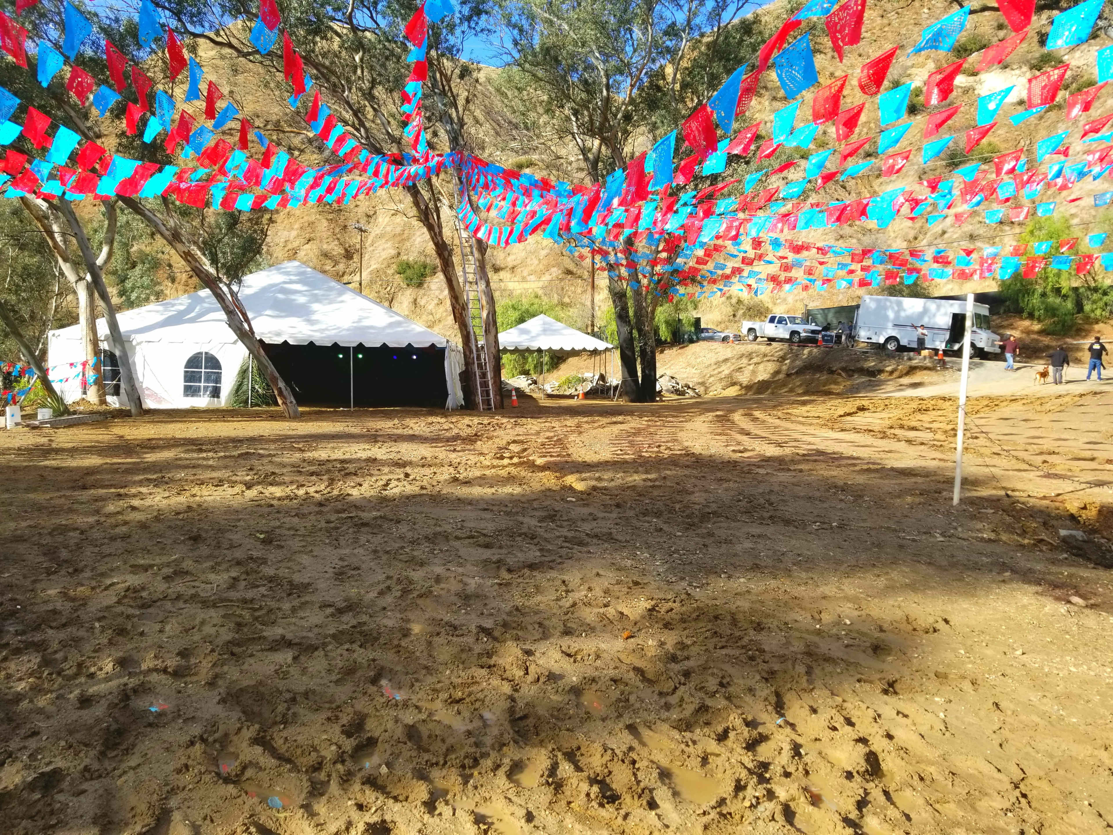 A large outdoor event space is decorated with colorful banners, featuring several tents in a sandy area surrounded by trees and hills.