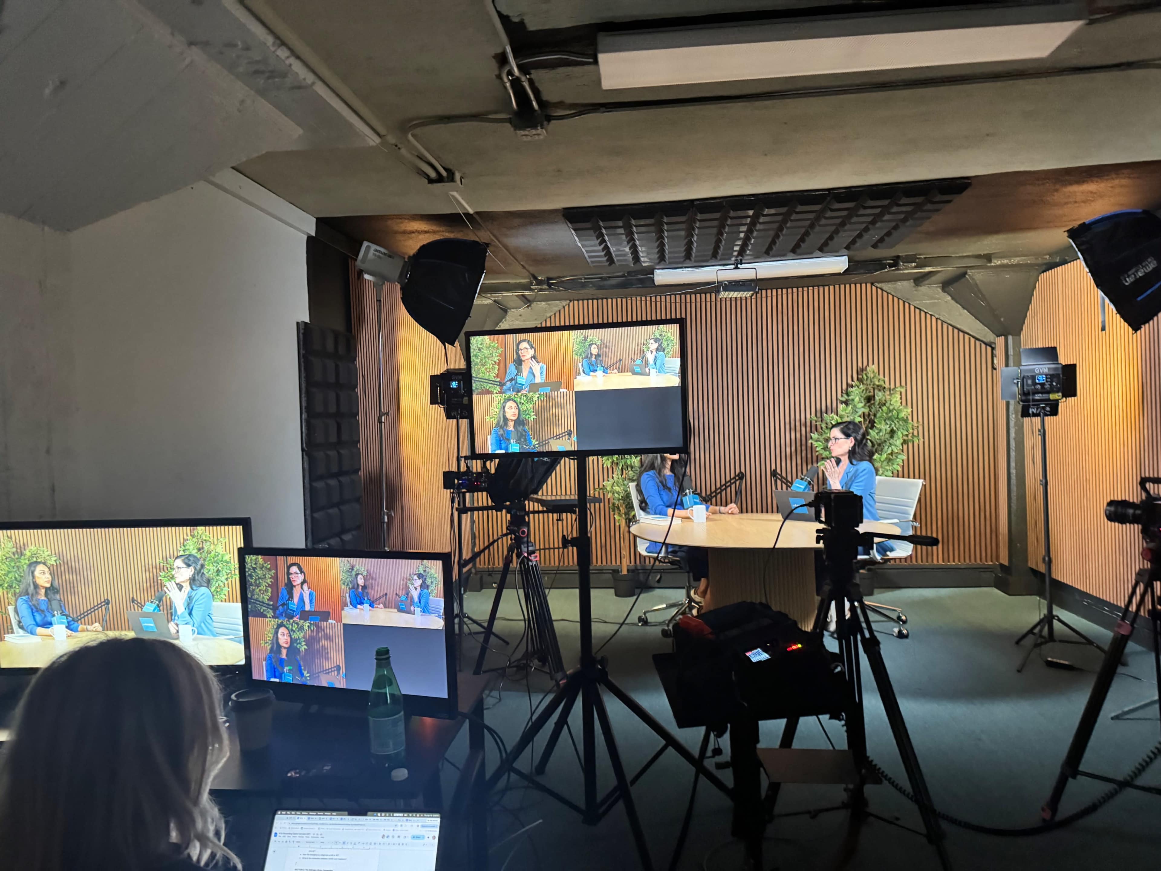 A video recording setup features two individuals discussing at a table, surrounded by cameras and lights, in a room with wooden paneling and greenery.