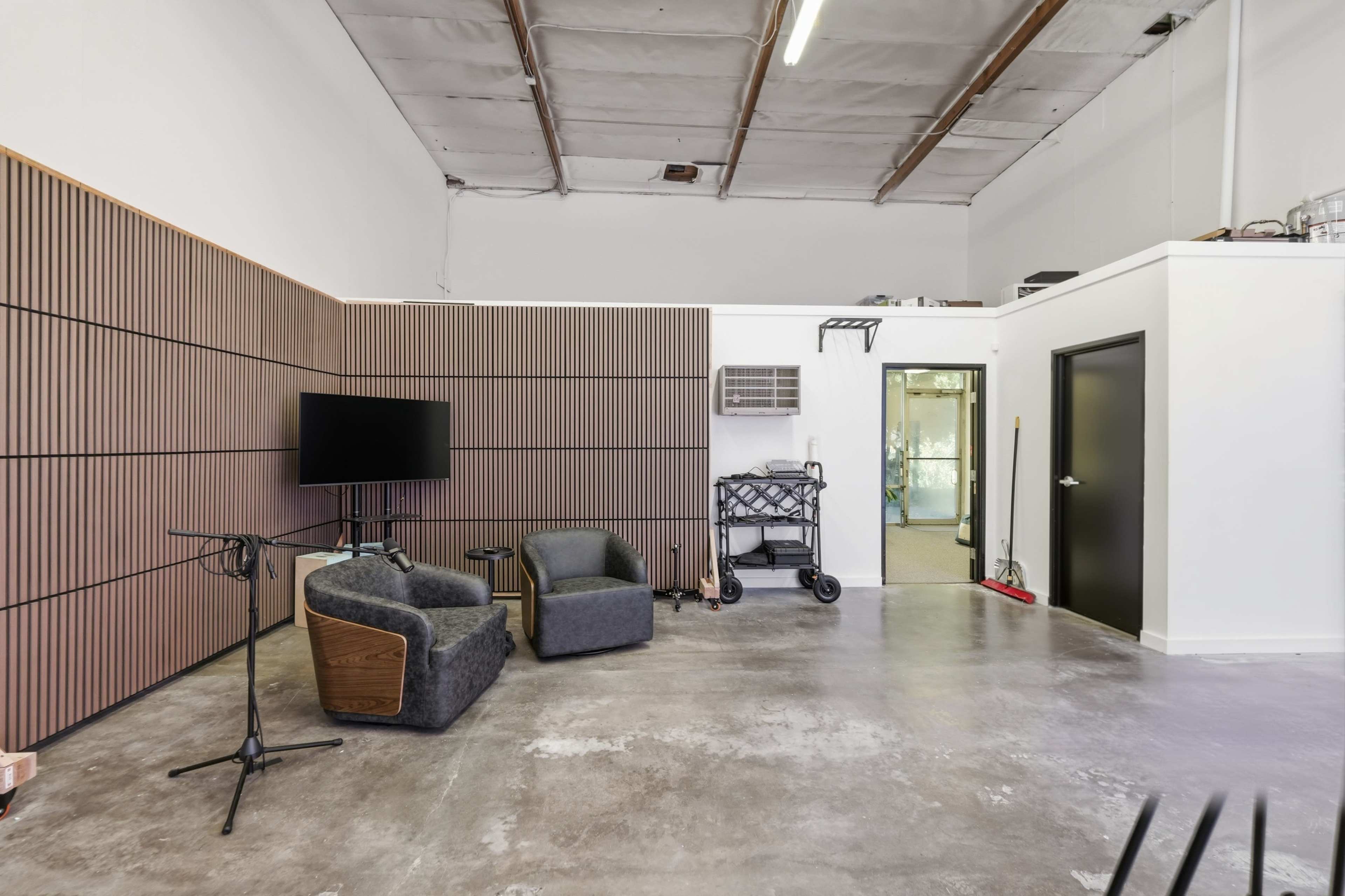 The image shows a spacious studio or rehearsal room featuring two gray chairs, a wall-mounted television, and a microphone stand, with a door leading to another area in the background.