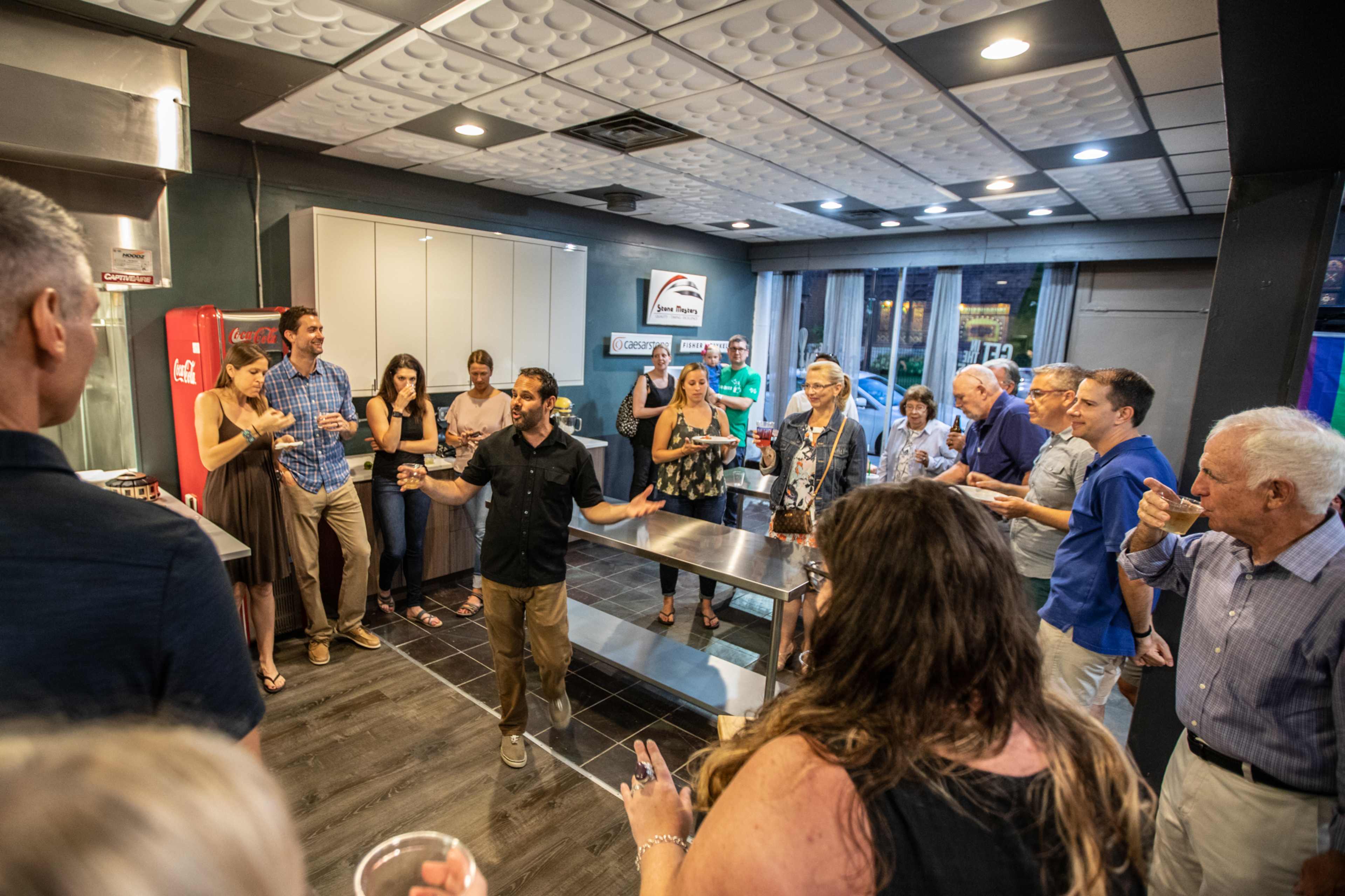 A group of people gathers in a modern kitchen space, with some standing around a central island while a man speaks in front of them.
