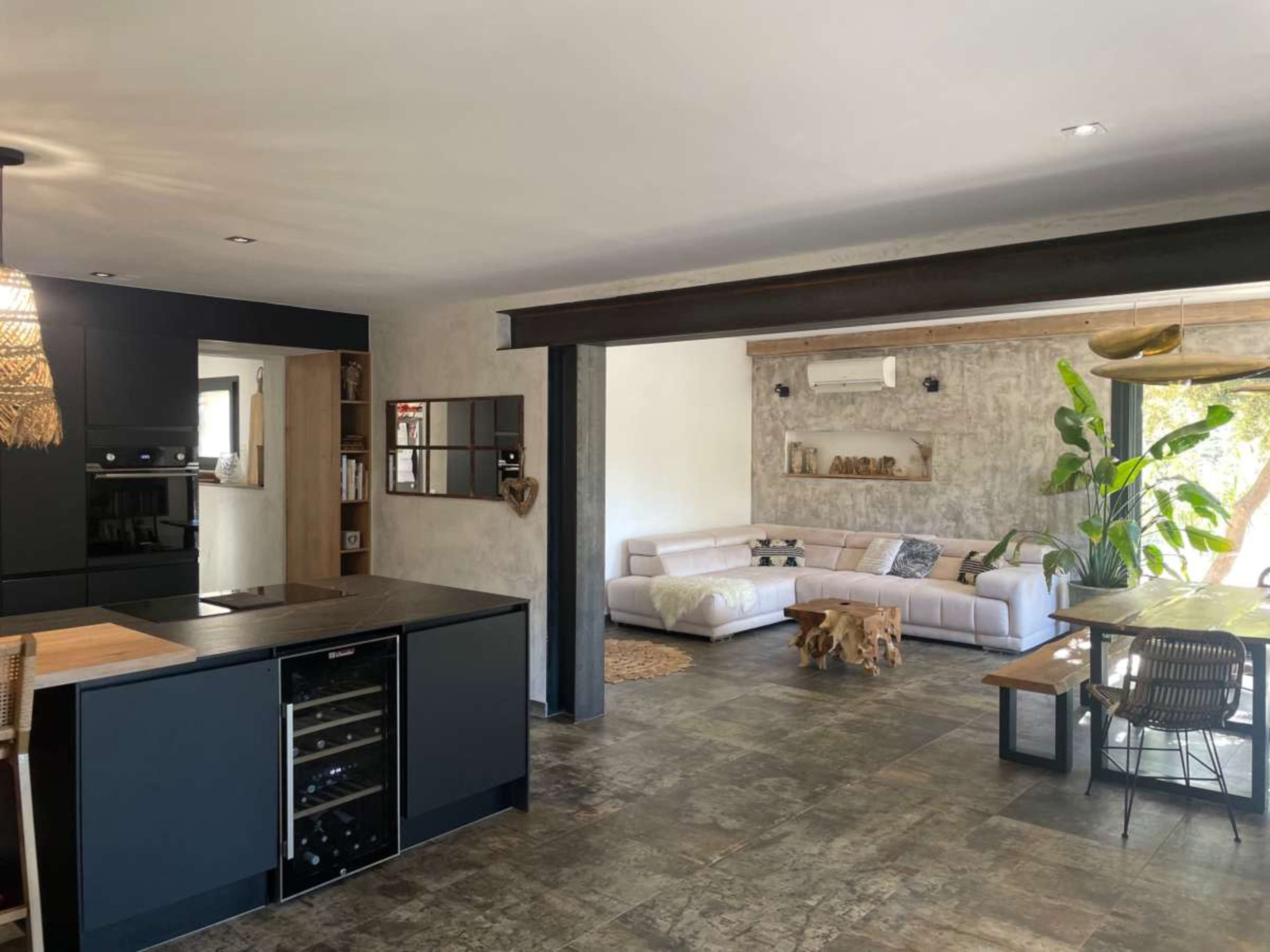 The image depicts a modern open-concept kitchen and living area, featuring sleek black cabinets, a central island, and a cozy seating arrangement with a large window and indoor plants.