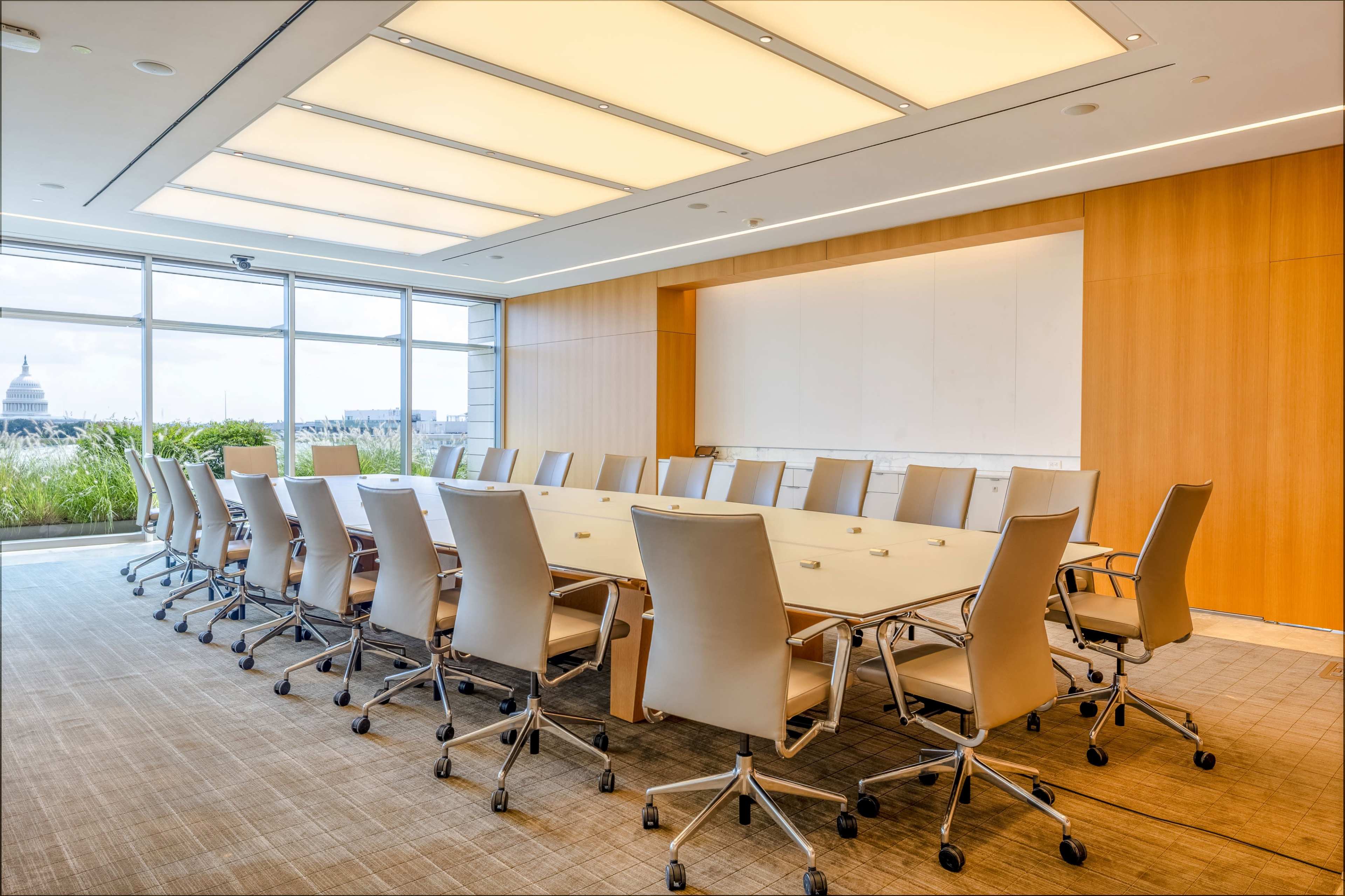A modern conference room features a long, rectangular table surrounded by ergonomic chairs, with large windows providing a view of the outside.