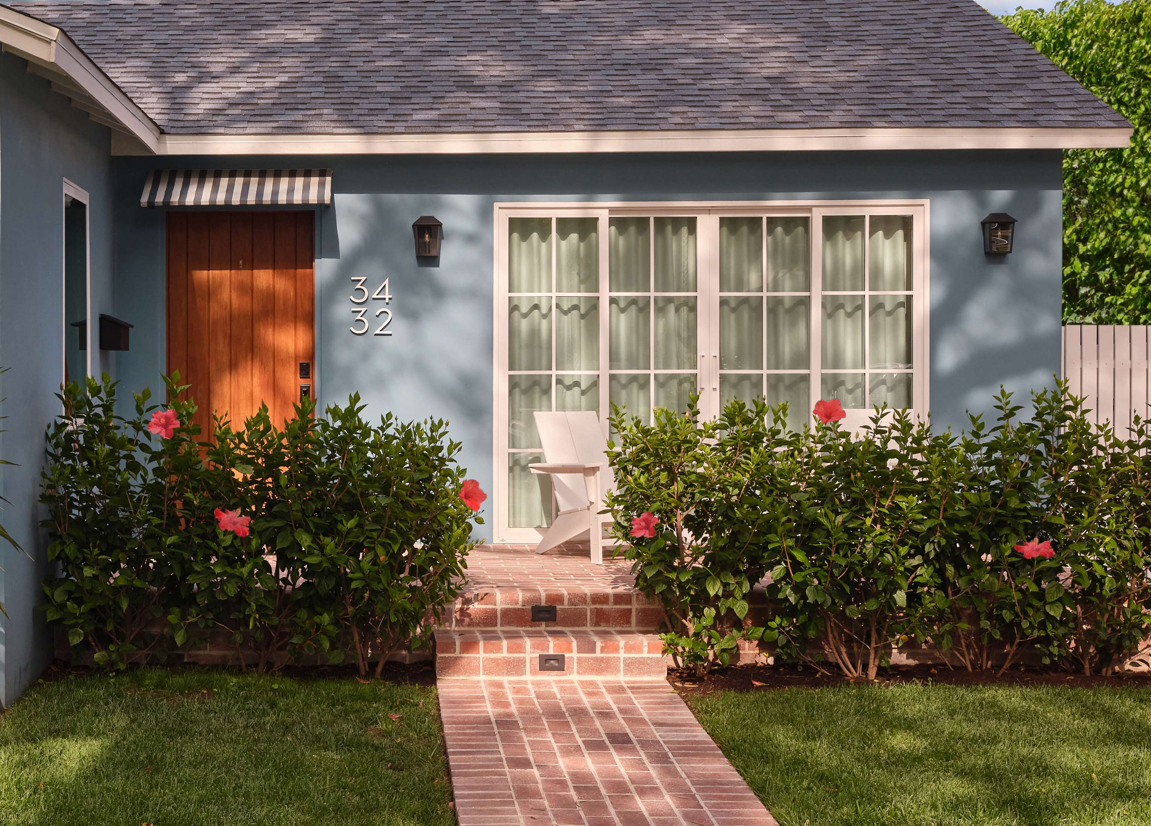 The image shows a blue house with a wooden door, white chairs on the porch, and a pathway lined with greenery and pink flowers.