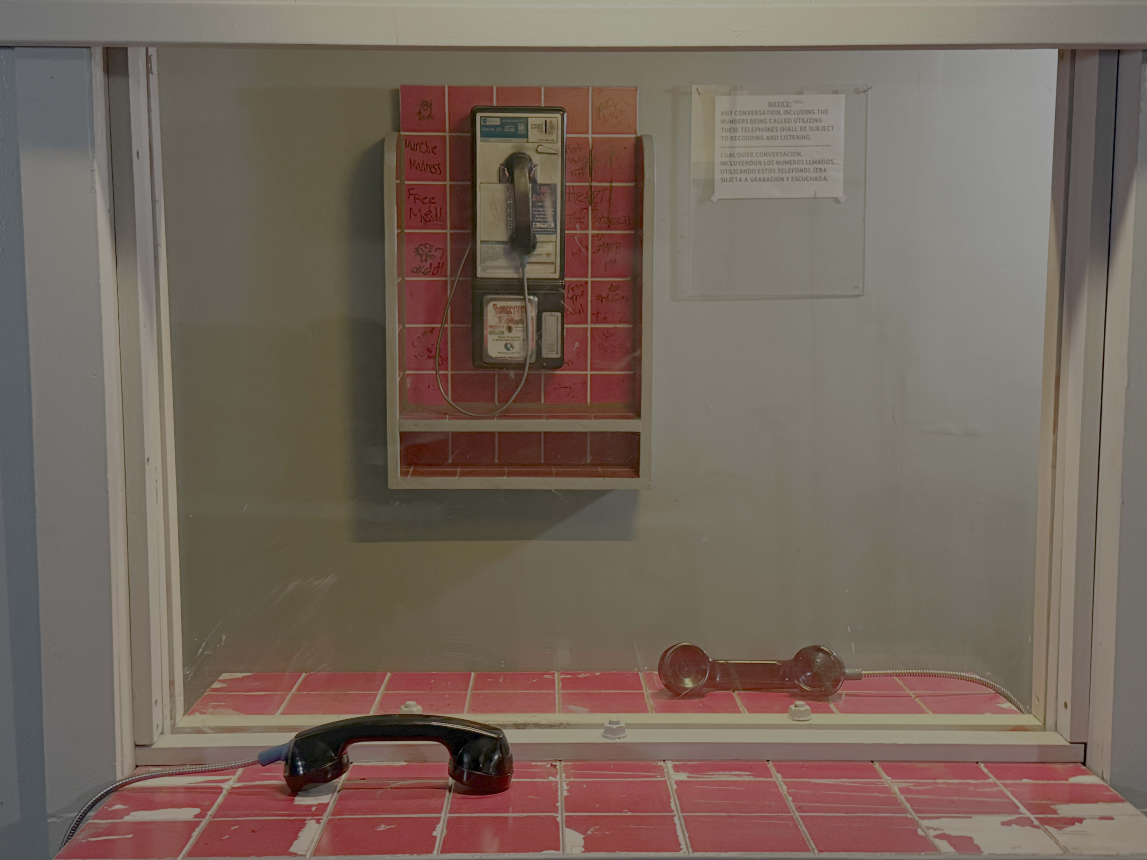 The image shows a clear phone booth with a payphone mounted on the wall and a disconnected handset resting on a red-tiled counter.