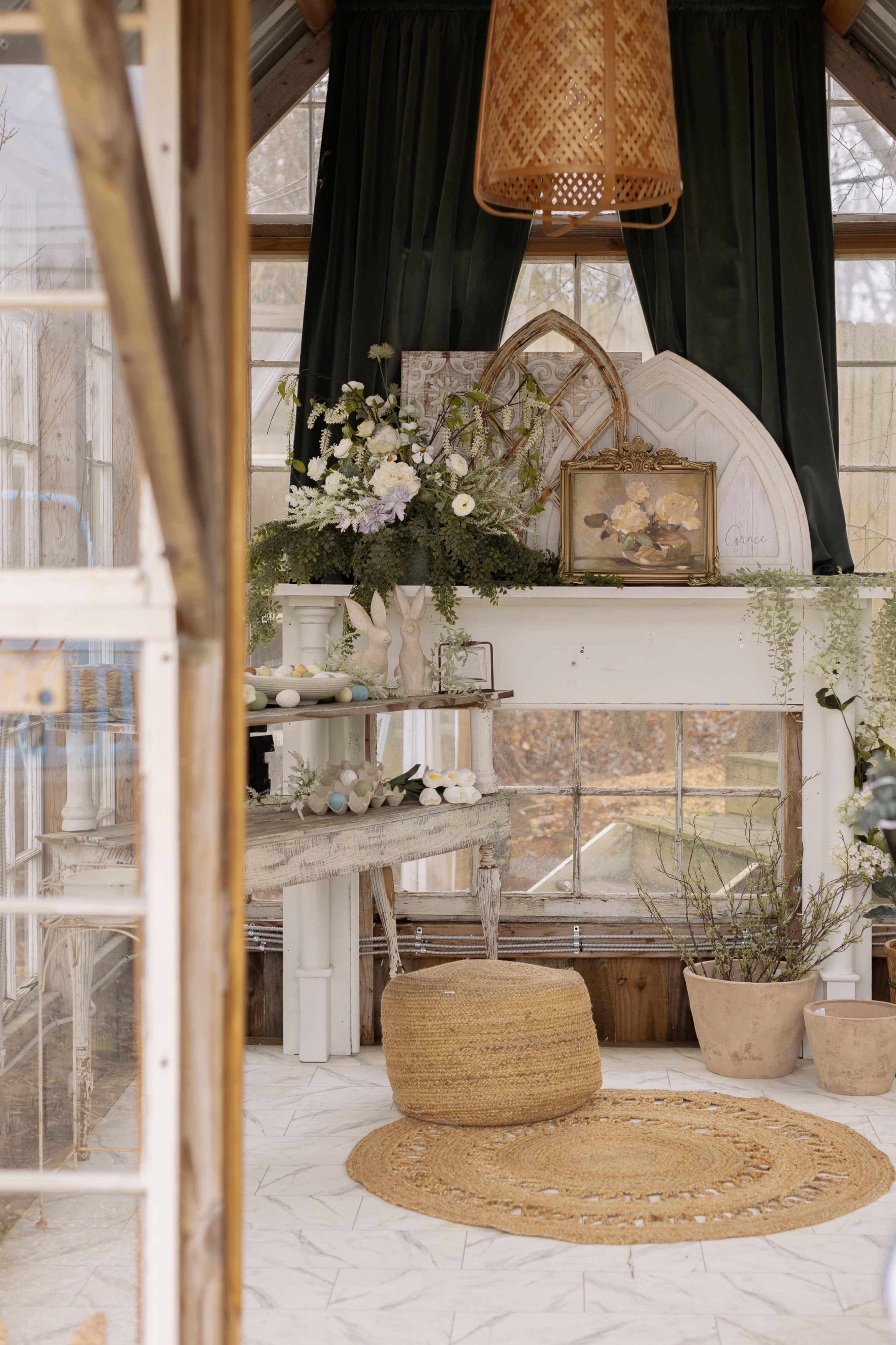 The image shows a cozy greenhouse interior with a decorative shelf featuring floral arrangements, a framed painting, and a patterned rug on the floor.