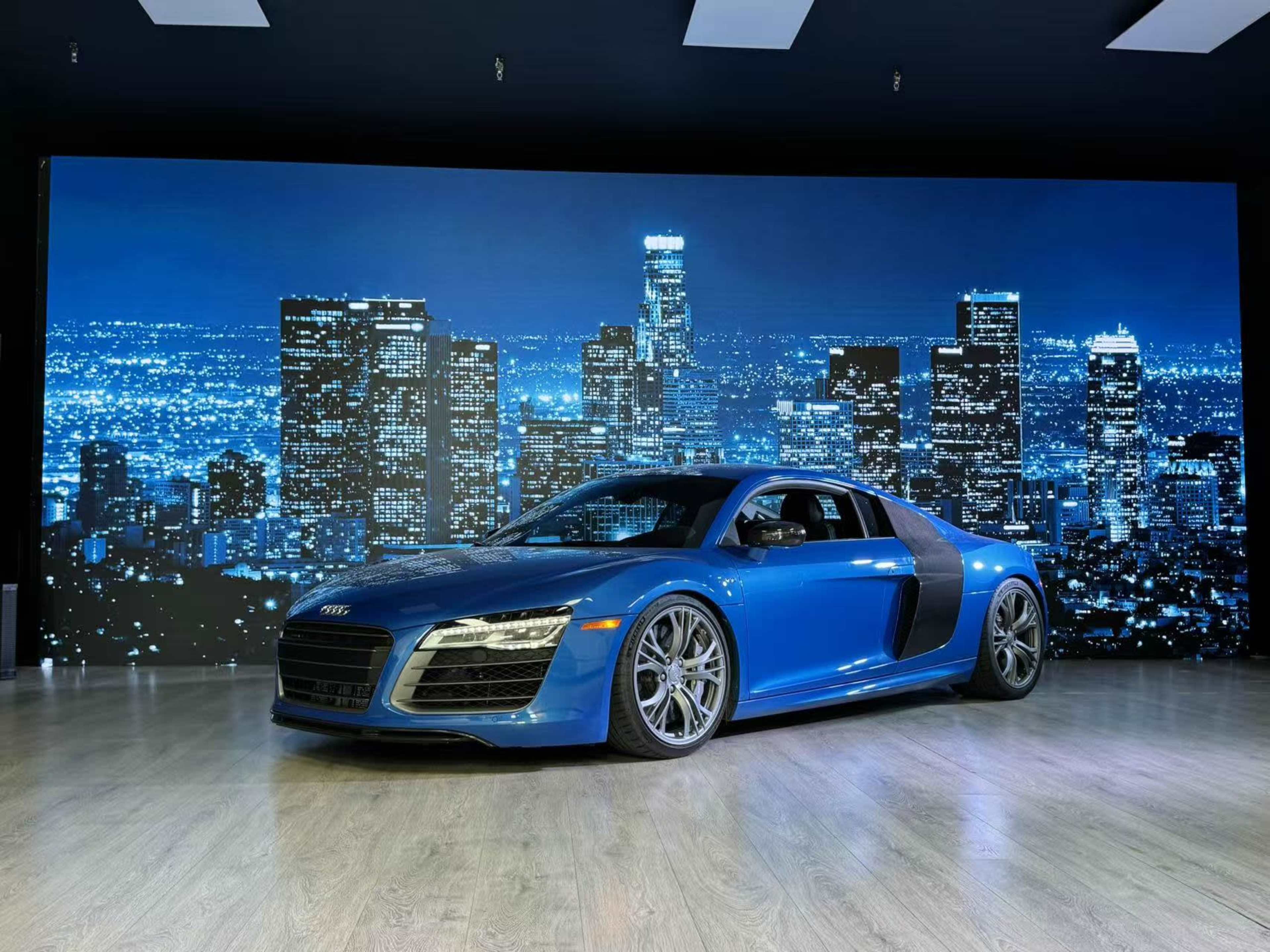 A blue Audi R8 is parked in front of a backdrop featuring a city skyline at night.