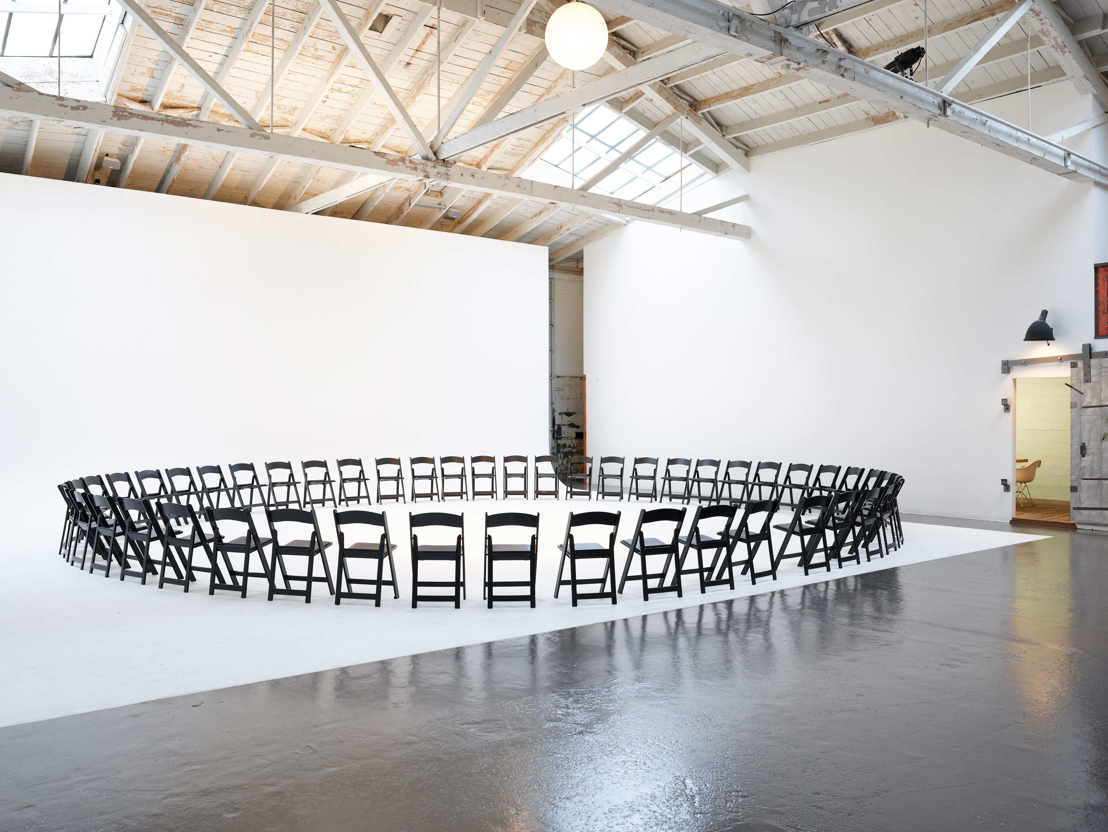 A circle of black chairs is arranged in an empty, well-lit space with white walls and a skylight.