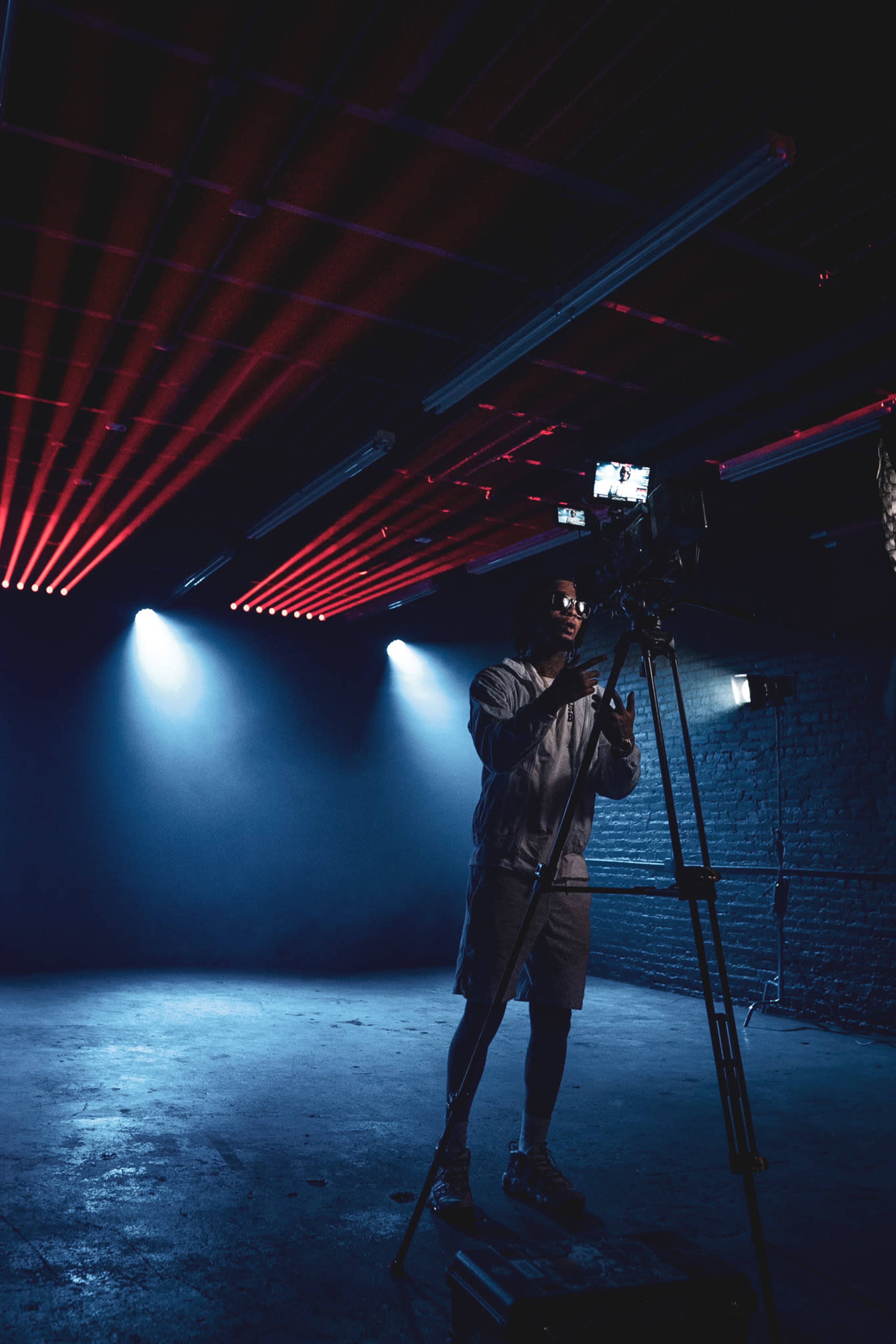 A person operates a camera on a tripod in a dimly lit space with beams of red and blue lights.