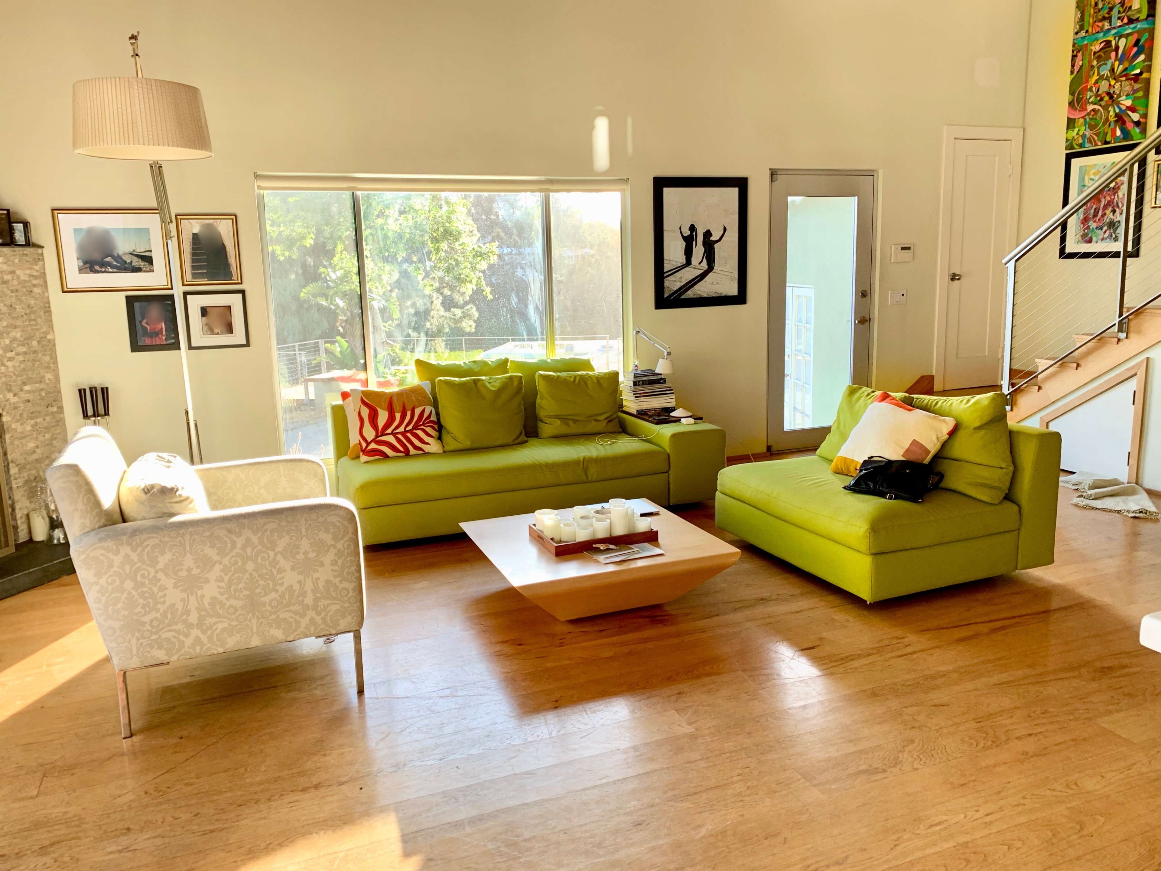 The image shows a brightly lit modern living room featuring green sofas, a wooden coffee table, and large windows that provide a view of the outdoors.