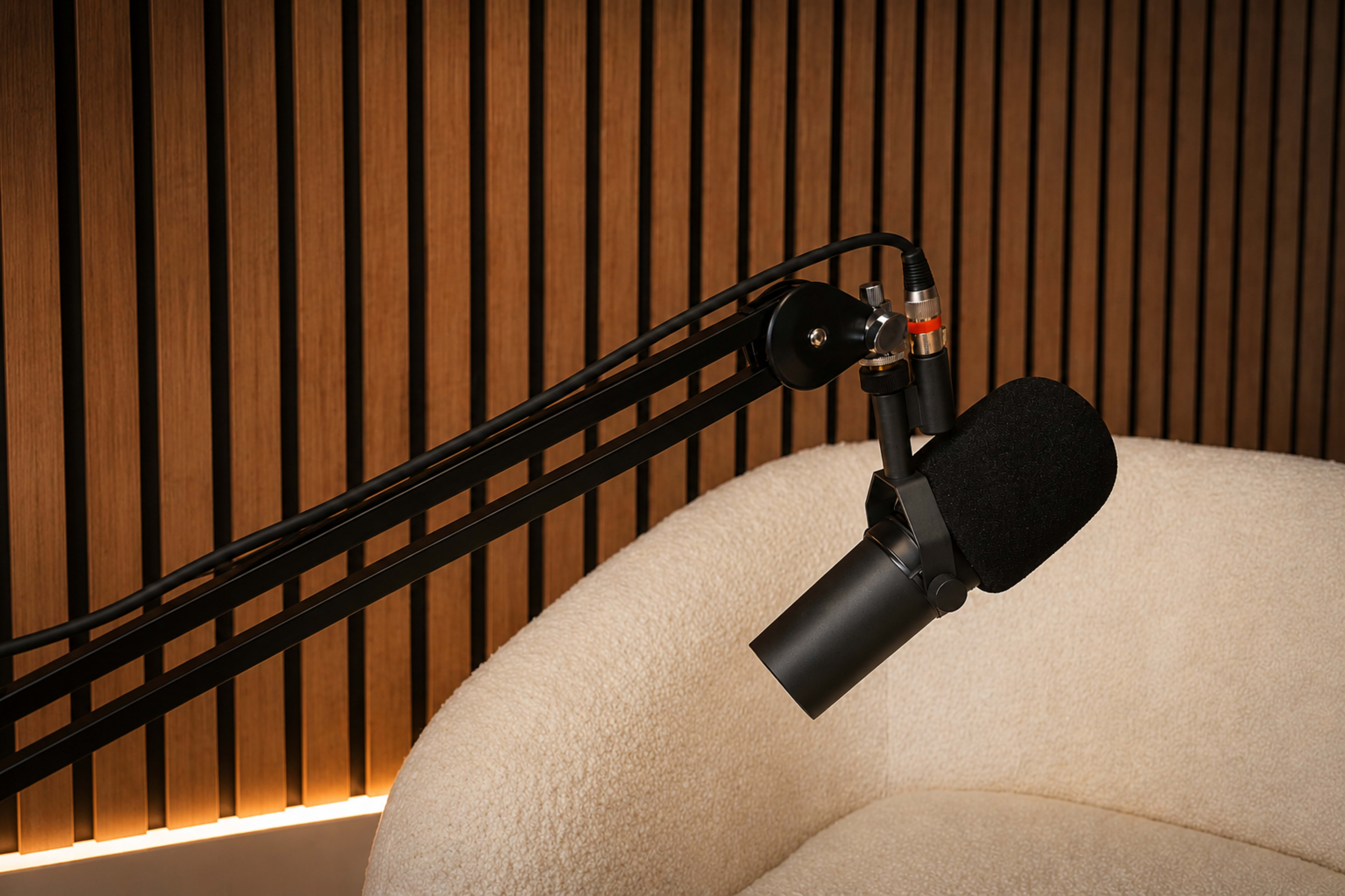 A microphone is positioned on a black adjustable arm next to a cream-colored cushioned chair, against a backdrop of wooden slats.