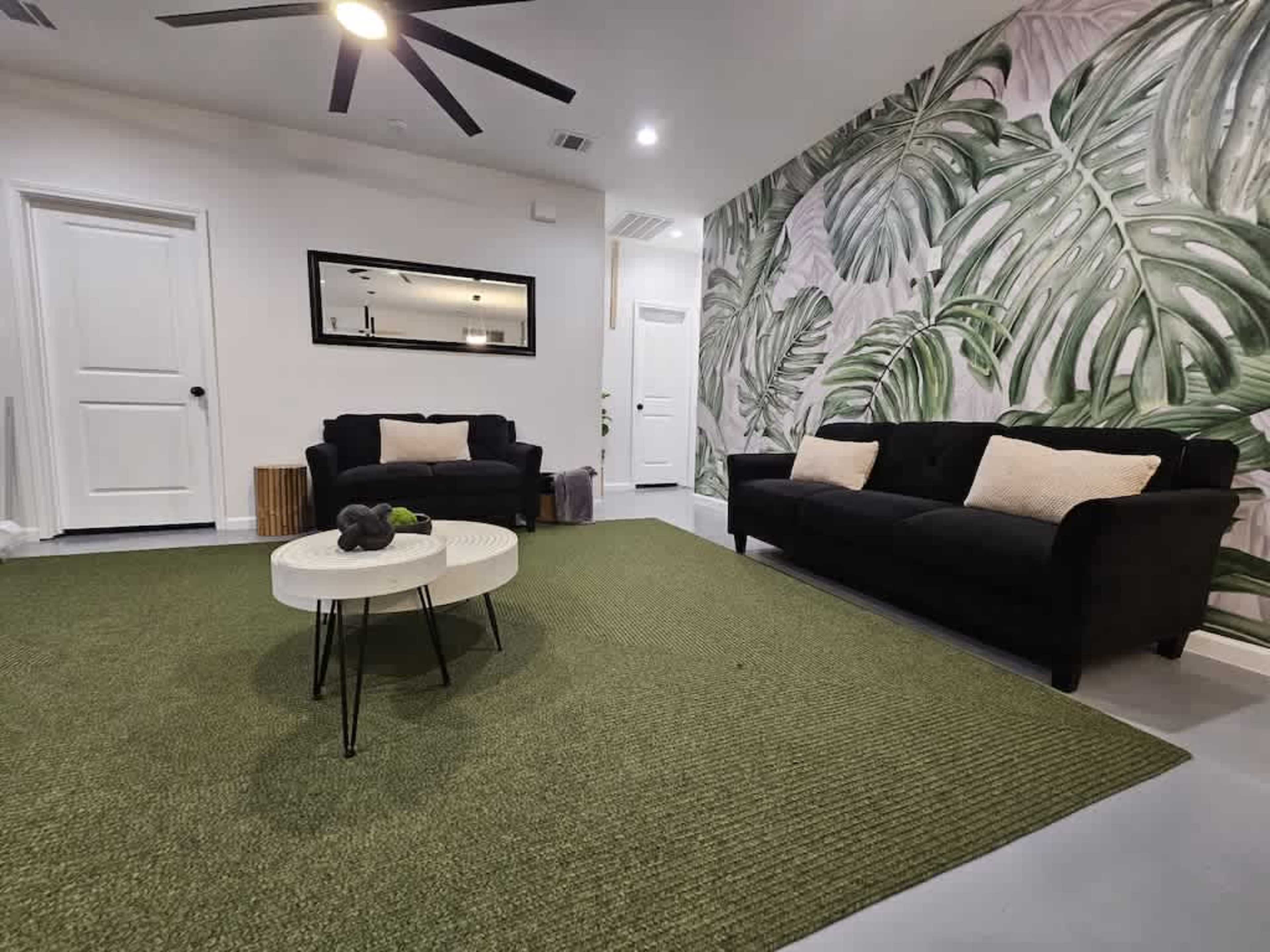 The living room features two black sofas, a round white coffee table, and a large green area rug, all against a wall adorned with a tropical leaf mural.