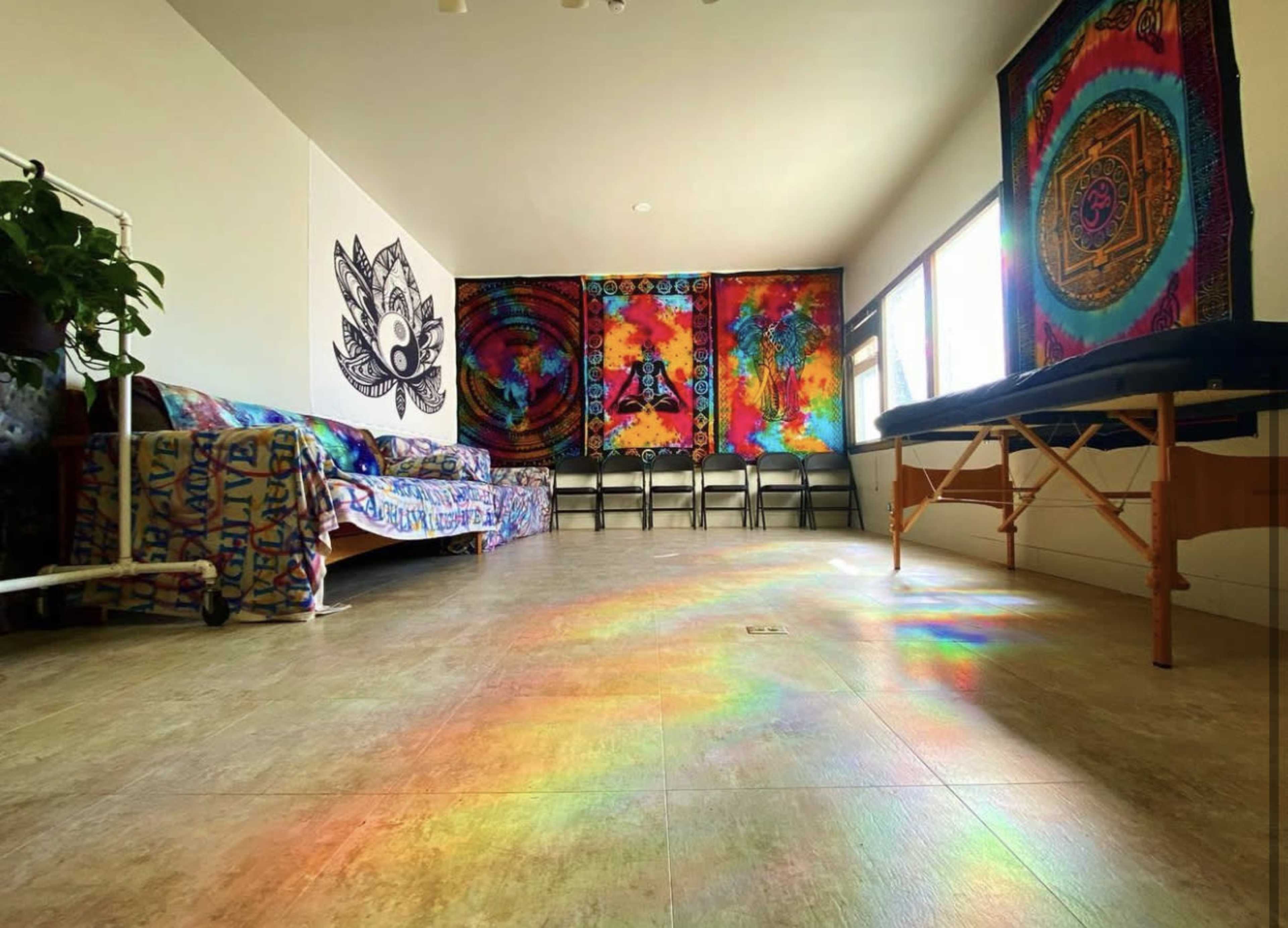 The interior of a brightly colored room features a patterned couch, folding chairs, and vibrant tapestries on the walls, with sunlight creating a rainbow effect on the floor.