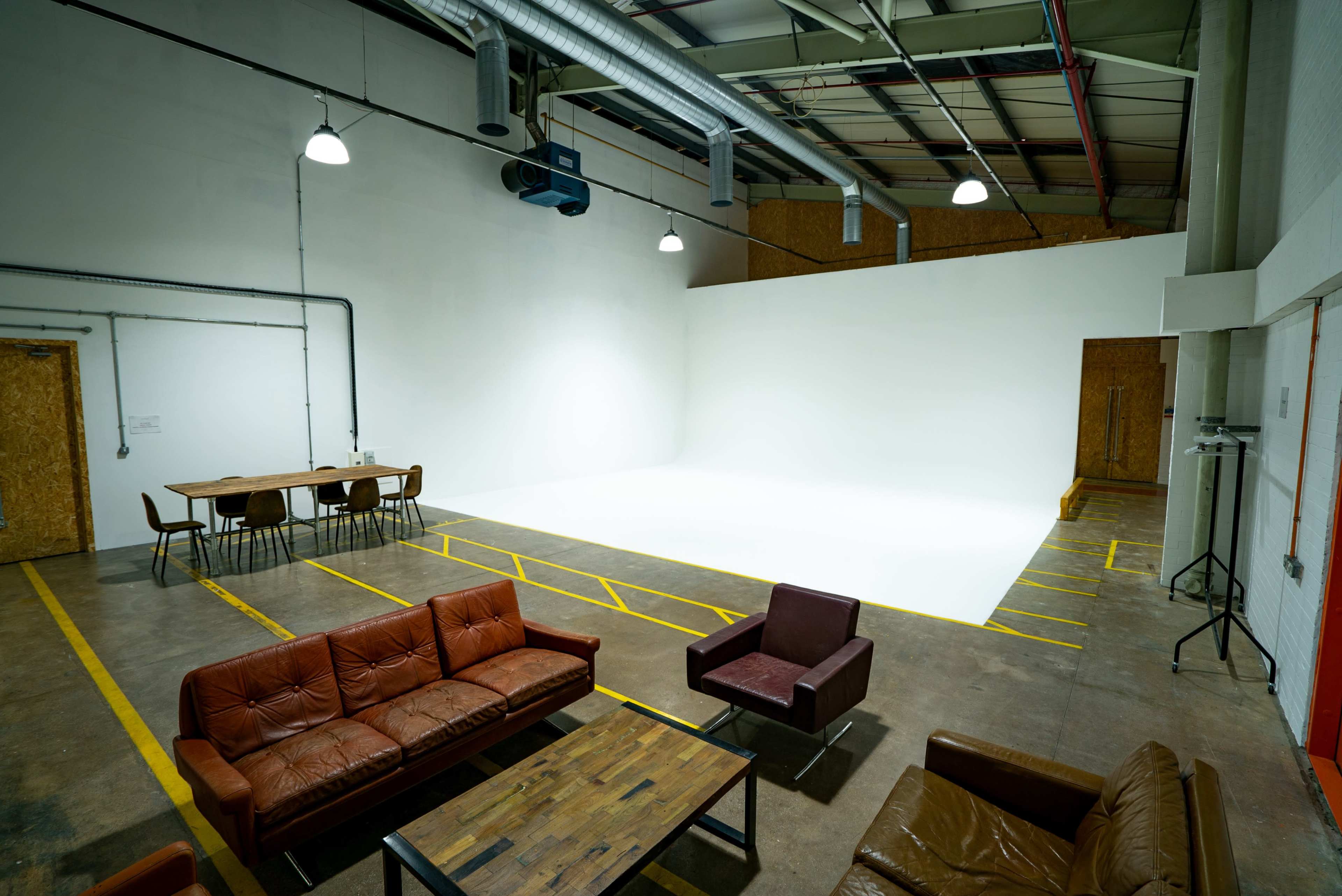 The image shows a spacious, empty studio with a large white backdrop, a seating area featuring brown leather couches, and a wooden meeting table with chairs.