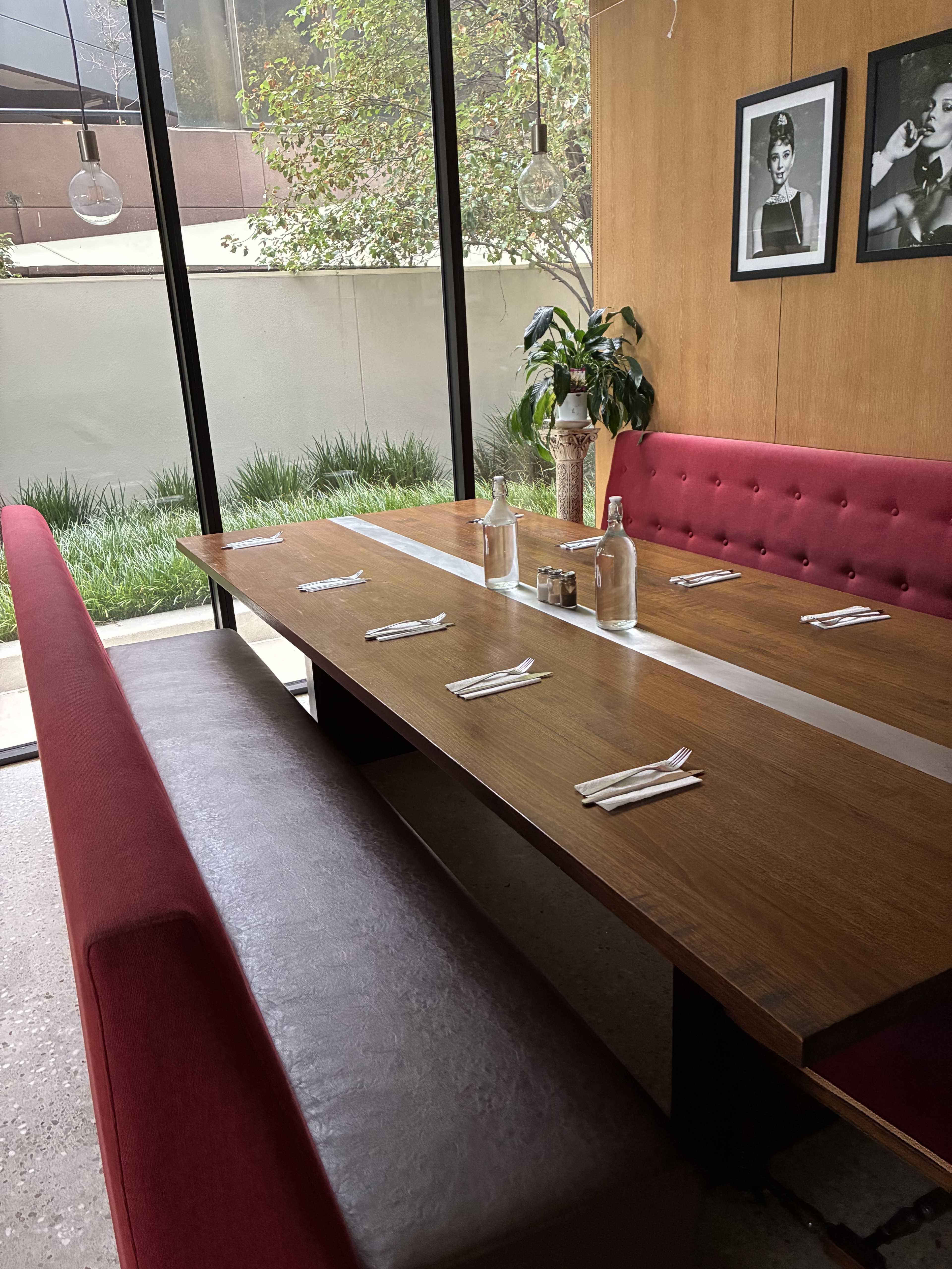 The image shows a modern restaurant interior featuring a large wooden table with red upholstered seating along one side and a decorative plant beside a window.
