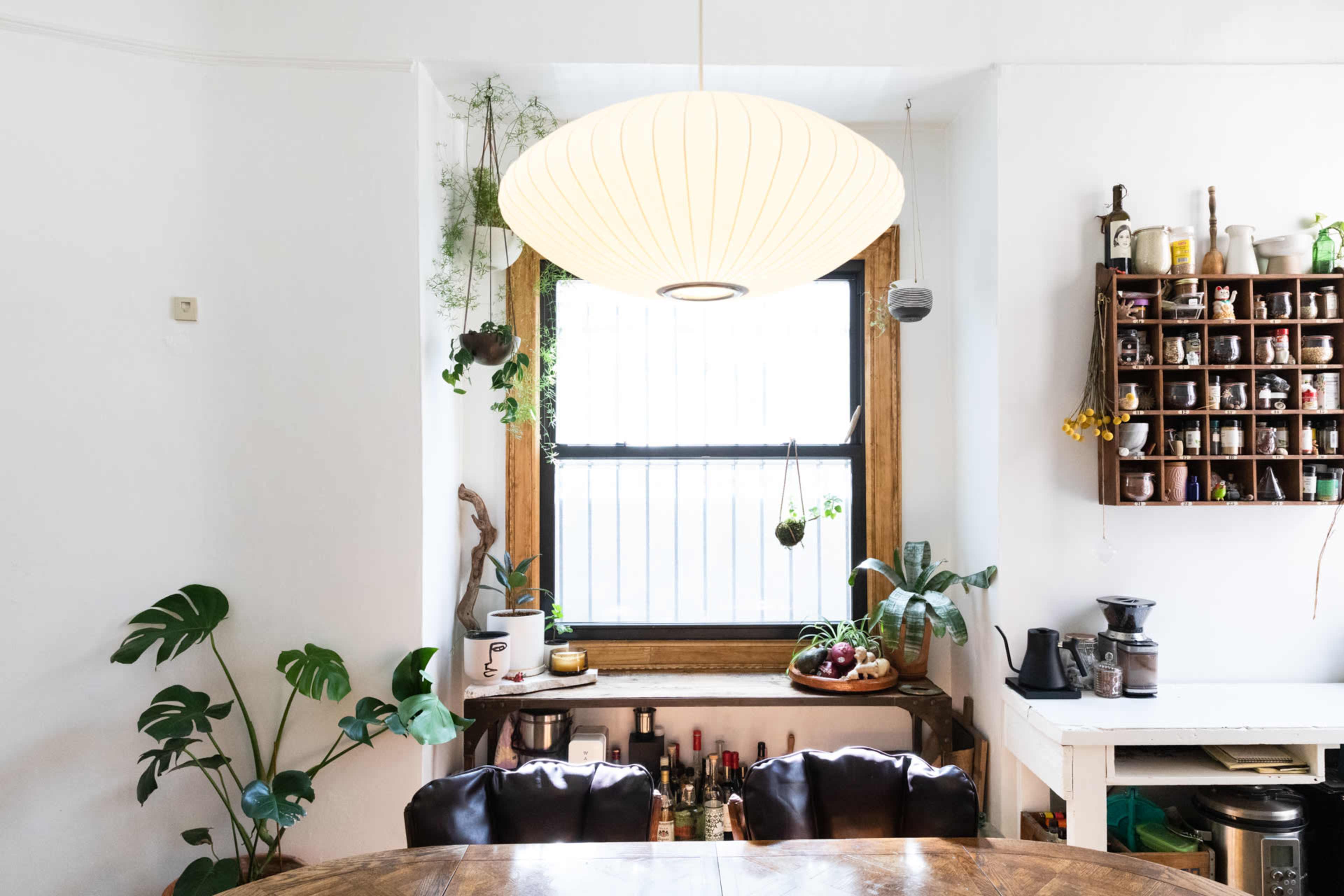 A cozy dining area features a round wooden table, potted plants, a large pendant light, and shelves filled with jars and kitchen items against a white wall.