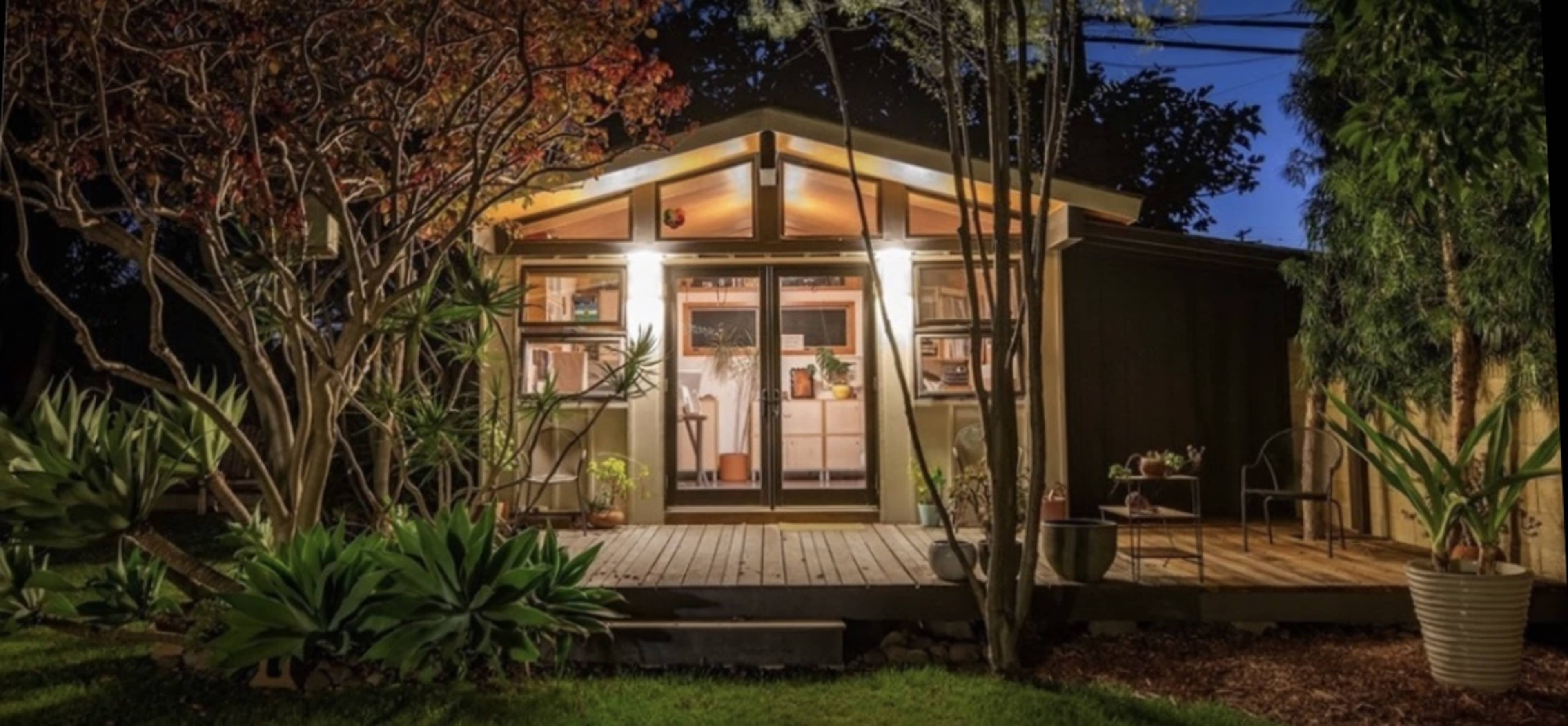 A modern wooden cabin with large windows is illuminated at night, surrounded by lush greenery and a wooden deck.