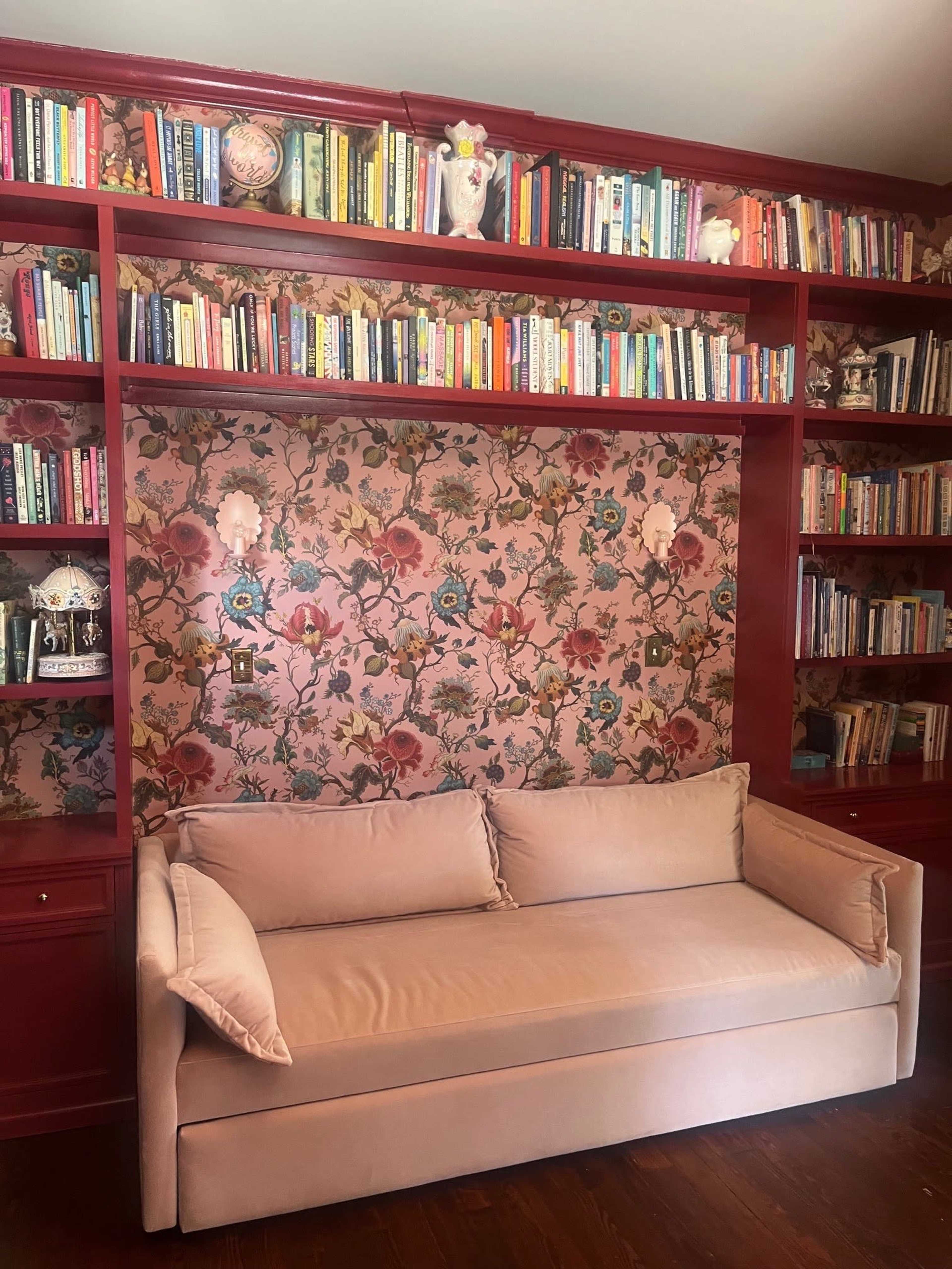 The image shows a pink velvet sofa in front of a patterned wallpaper and a bookshelf filled with various books.