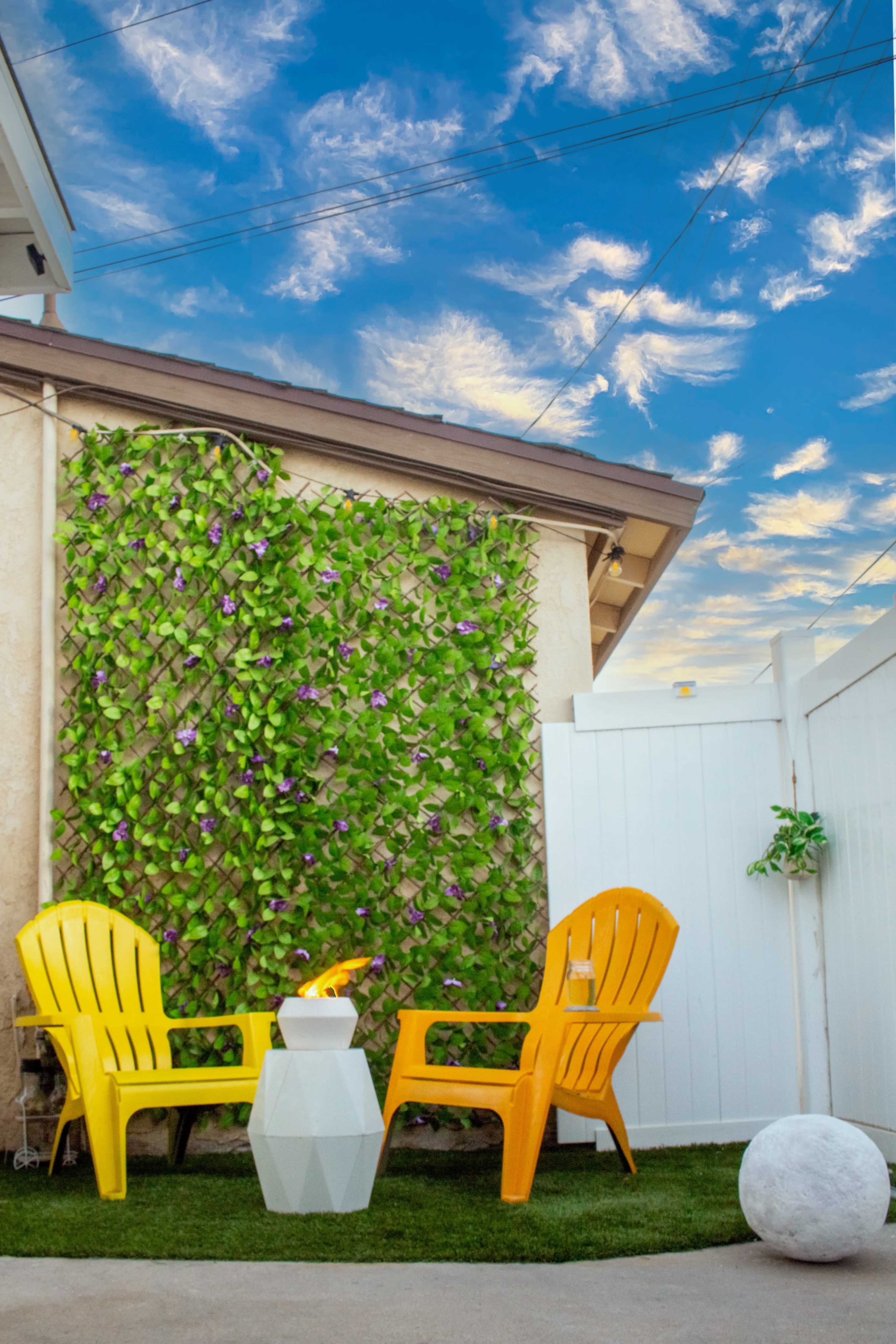 A small outdoor seating area features two yellow chairs and a geometric white table against a backdrop of a green wall and a blue sky with clouds.