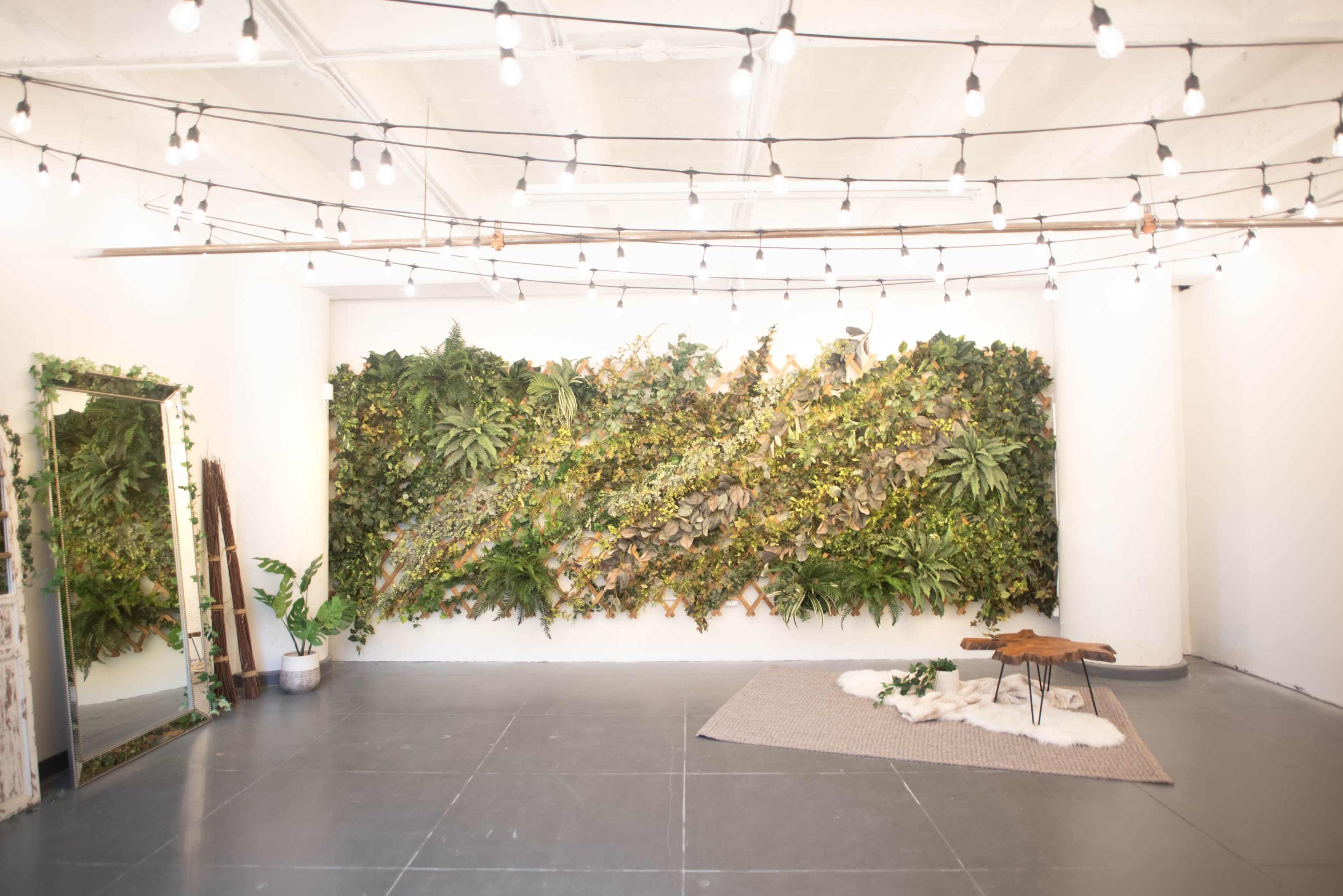 A bright, minimalist interior space featuring a wall of greenery, string lights overhead, and a simple wooden table with a small decorative arrangement on a textured rug.