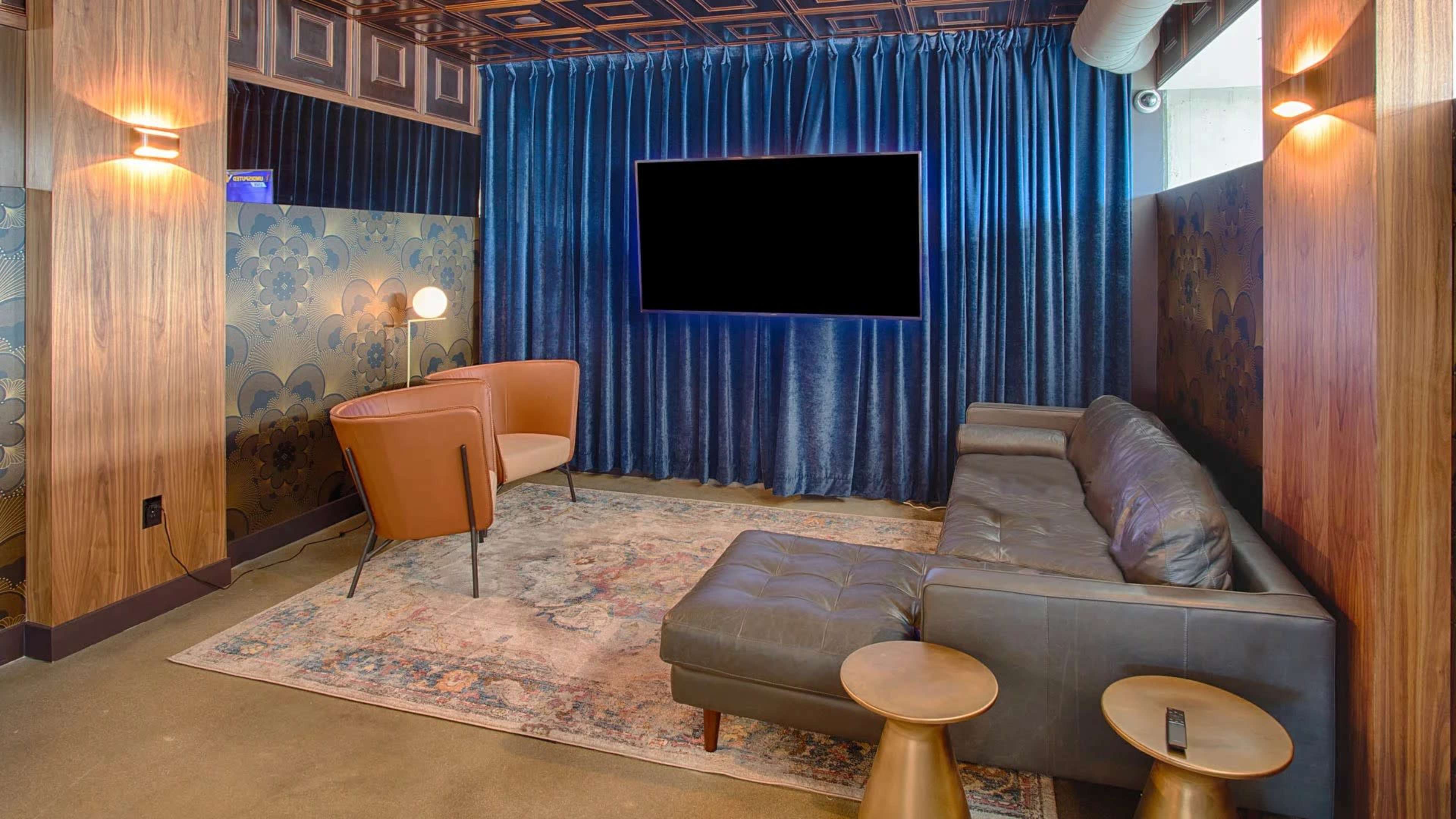 A stylish lounge area with a dark leather sofa, two chairs, a rug, and a large television mounted on the wall, all surrounded by blue curtains and wood paneling.