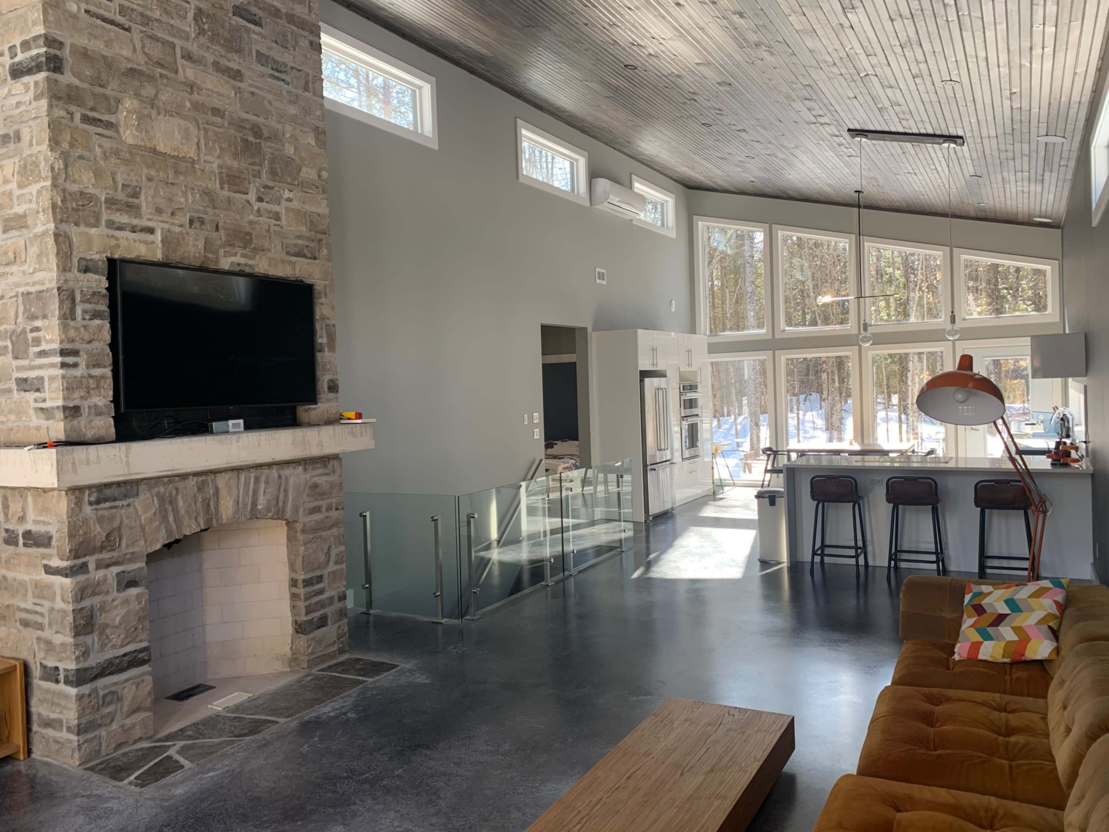 A modern living room featuring a stone fireplace, large windows with views of a wooded area, and a contemporary seating arrangement.