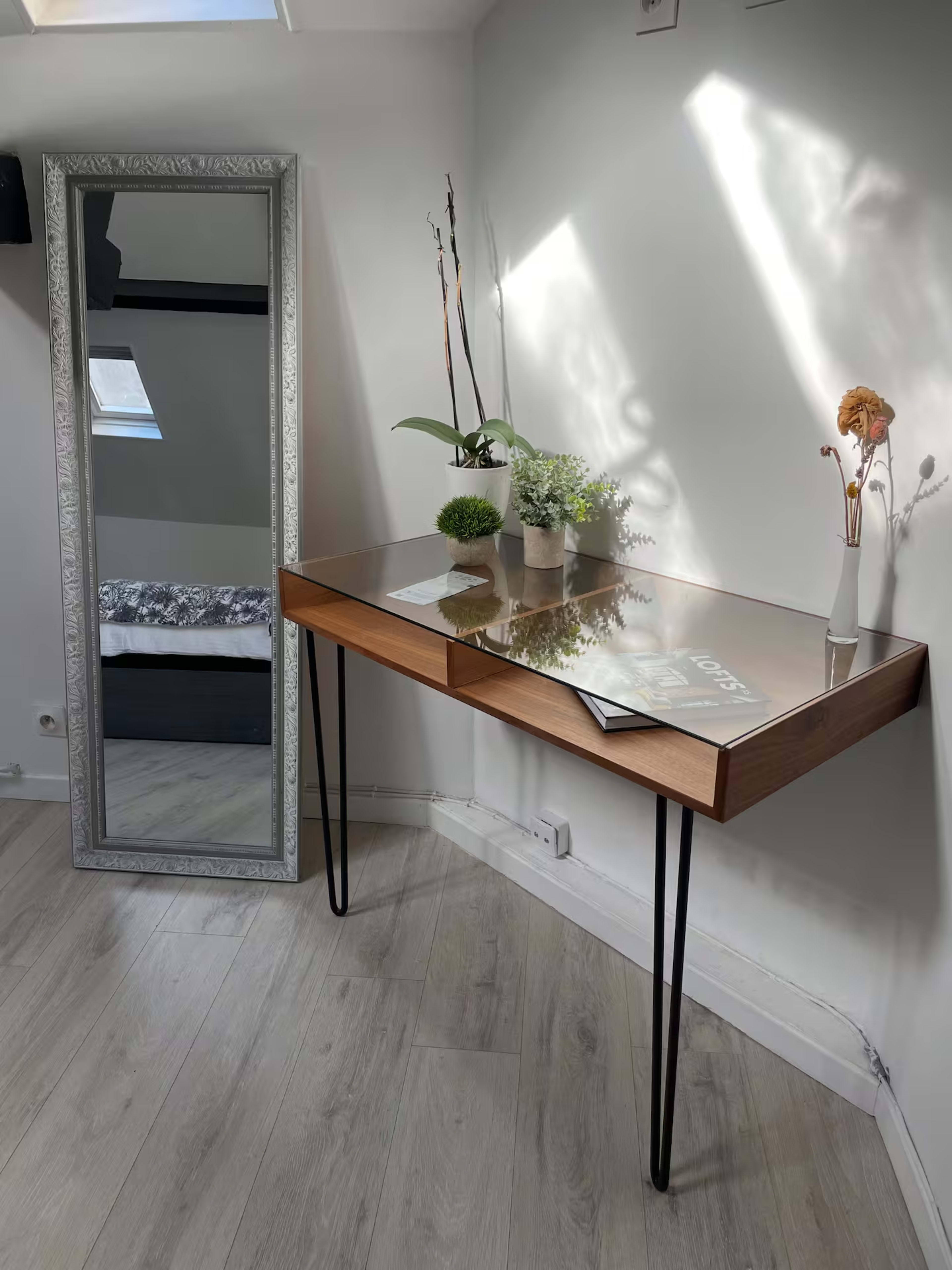 A wooden table with a glass top and two decorative plants stands against a wall, next to a tall mirror.