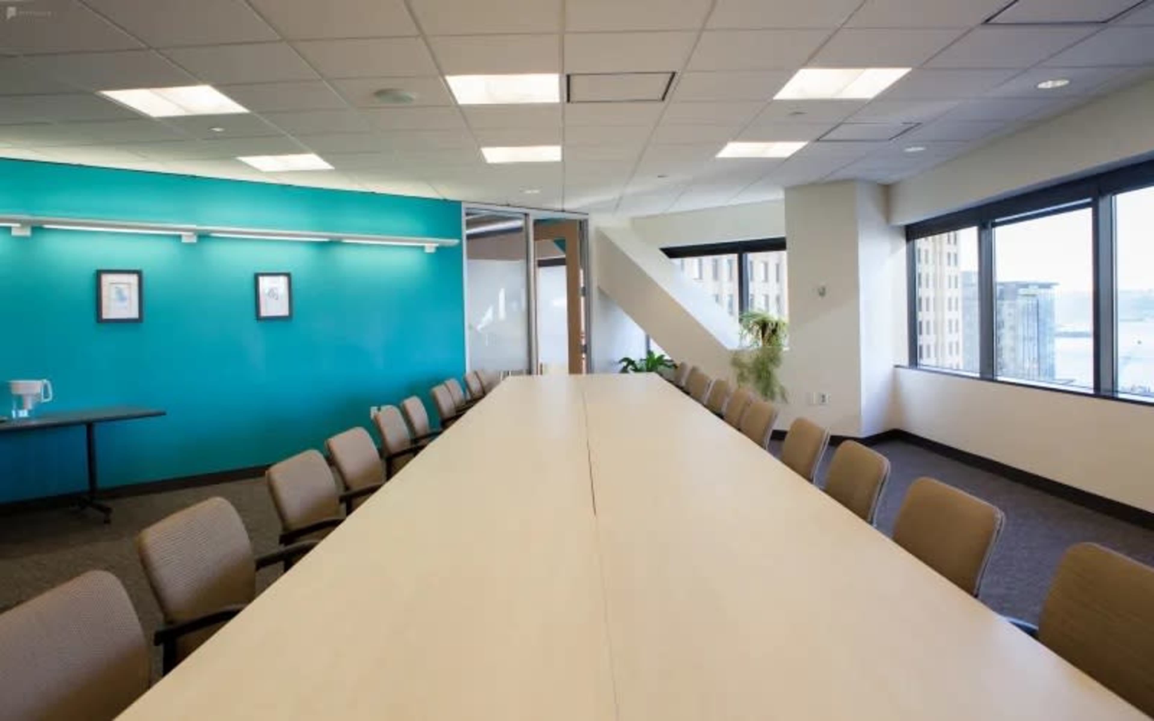 A long, white conference table is centered in a bright meeting room with teal walls and large windows offering a view of the city.