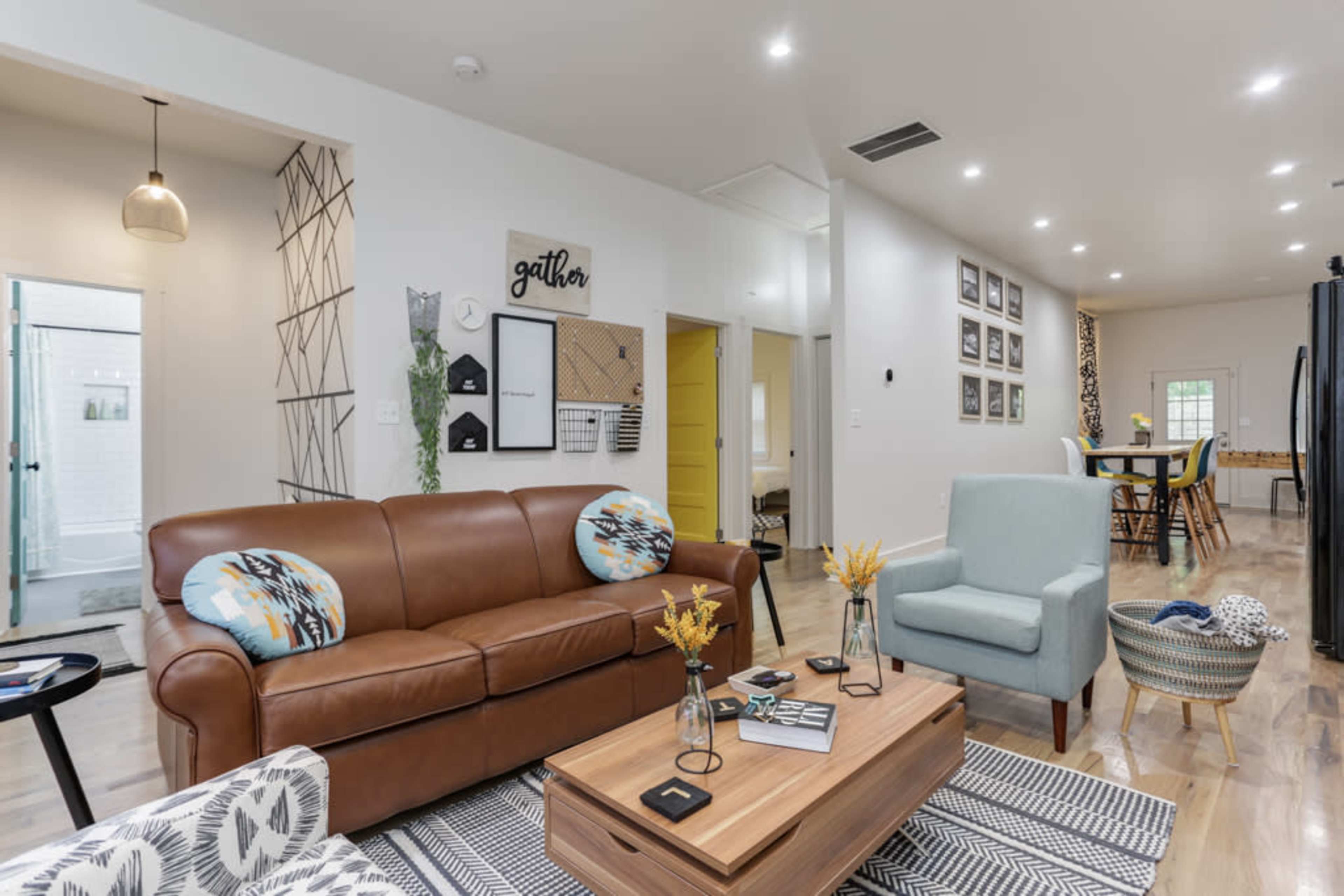A modern living space with a brown leather sofa, blue accent chairs, a wooden coffee table, and a dining area in the background featuring multiple framed pictures on the wall.