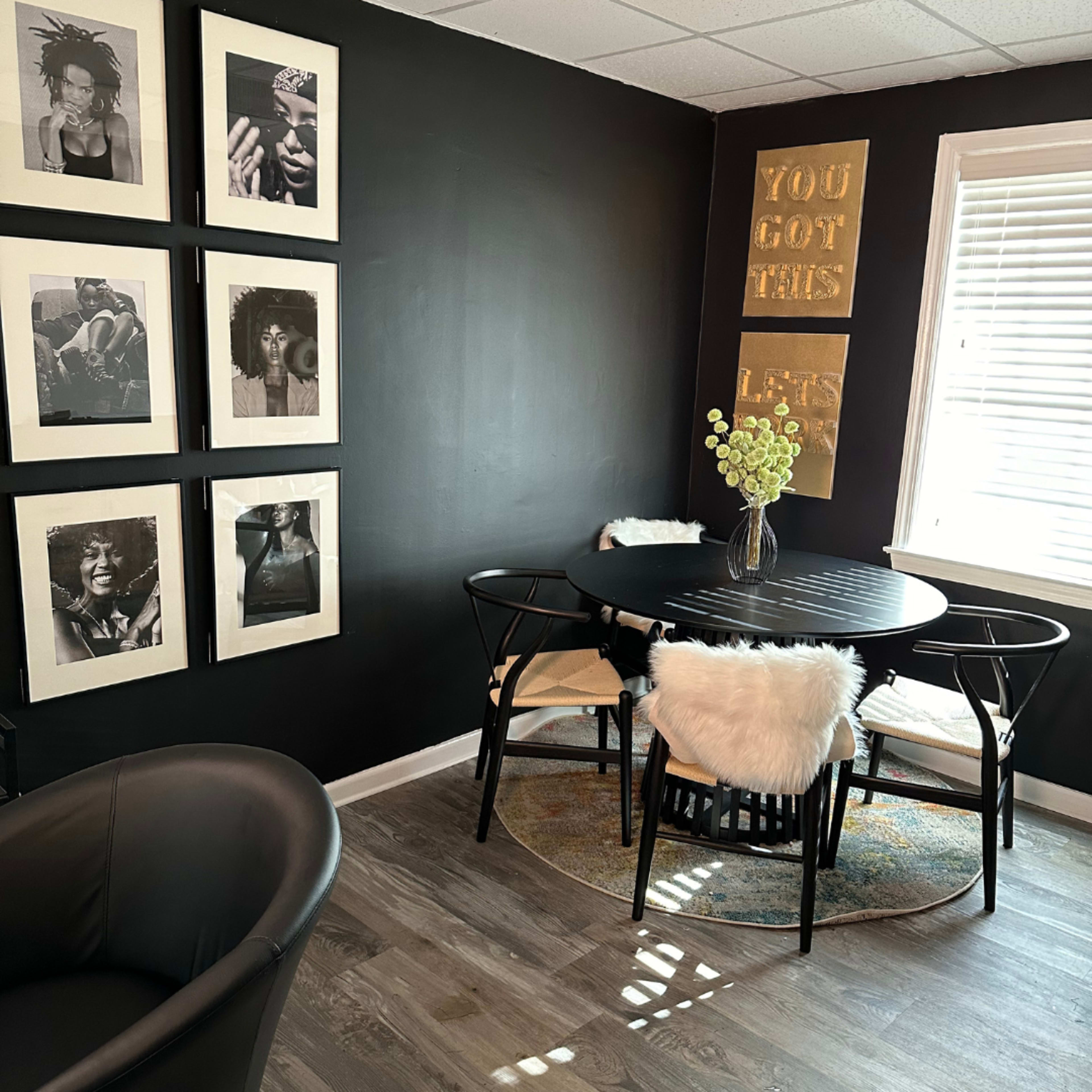 A black-walled room features a round black table surrounded by four chairs, adorned with cozy white seat covers and framed photographs on the wall.