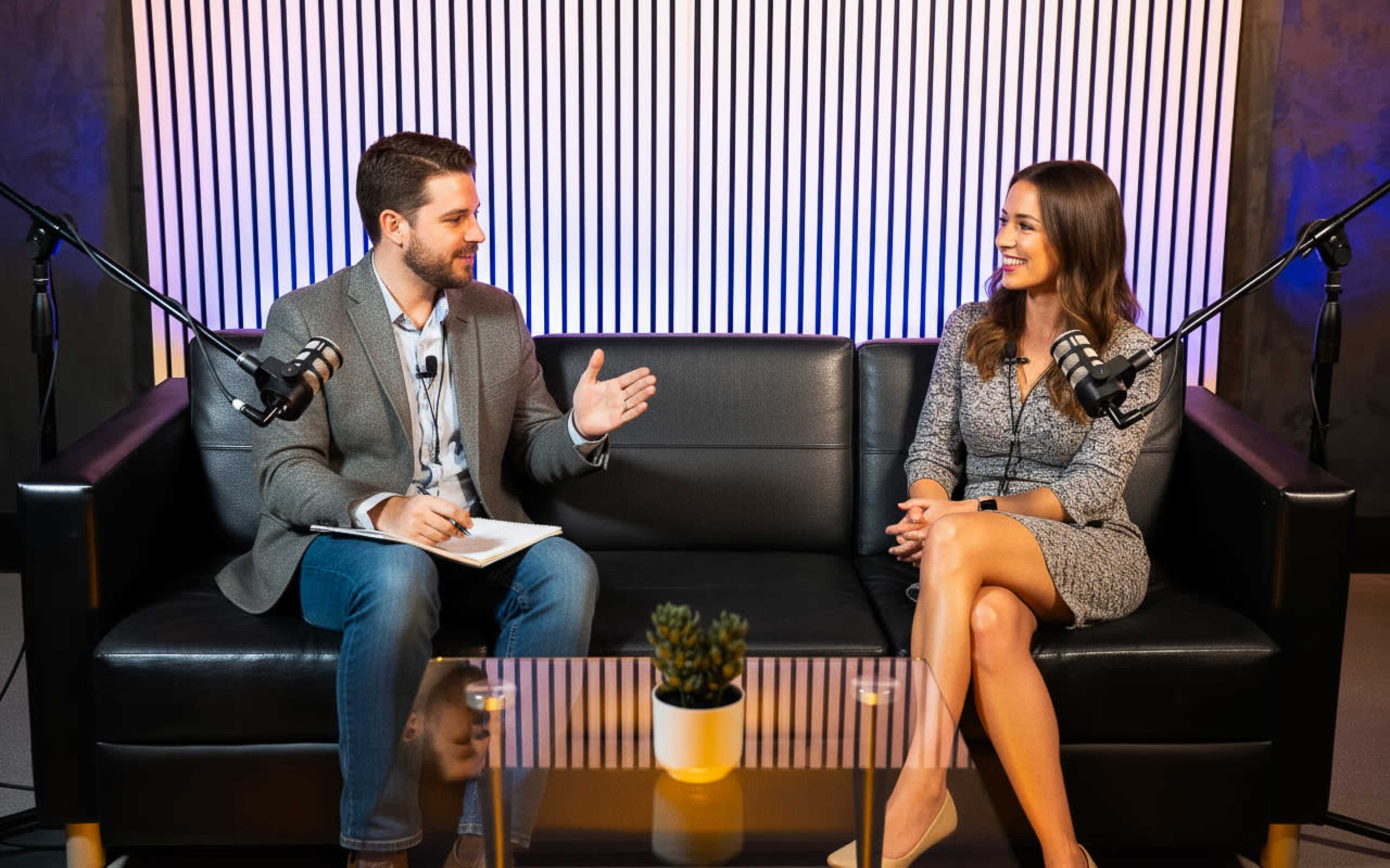 A man and a woman are sitting on a black couch in a studio, engaged in a conversation, with microphones in front of them and a small plant on a glass table between them.