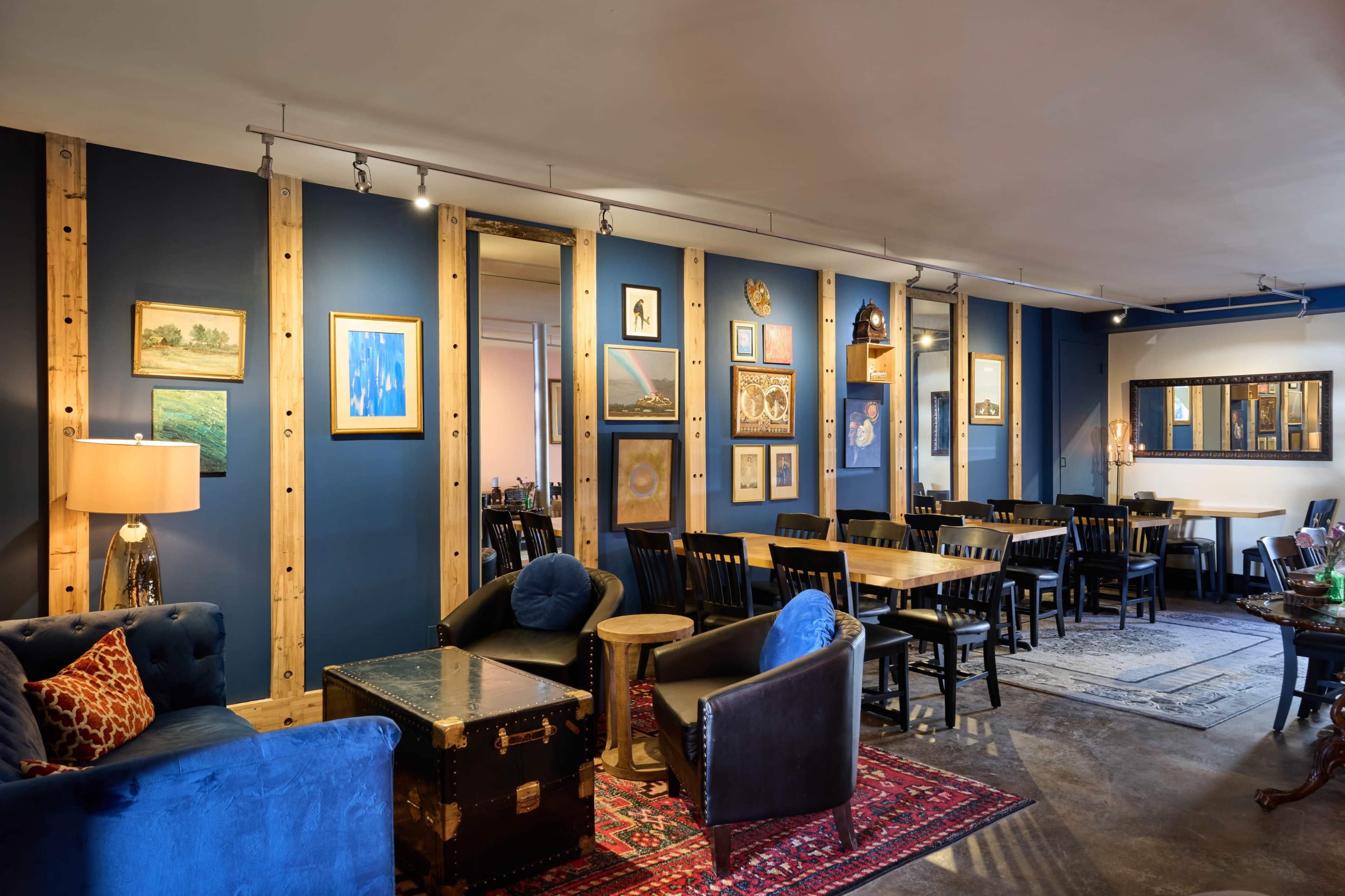 The image shows a cozy café with blue walls, wooden accents, and a mix of seating options including chairs and sofas, adorned with various artworks.