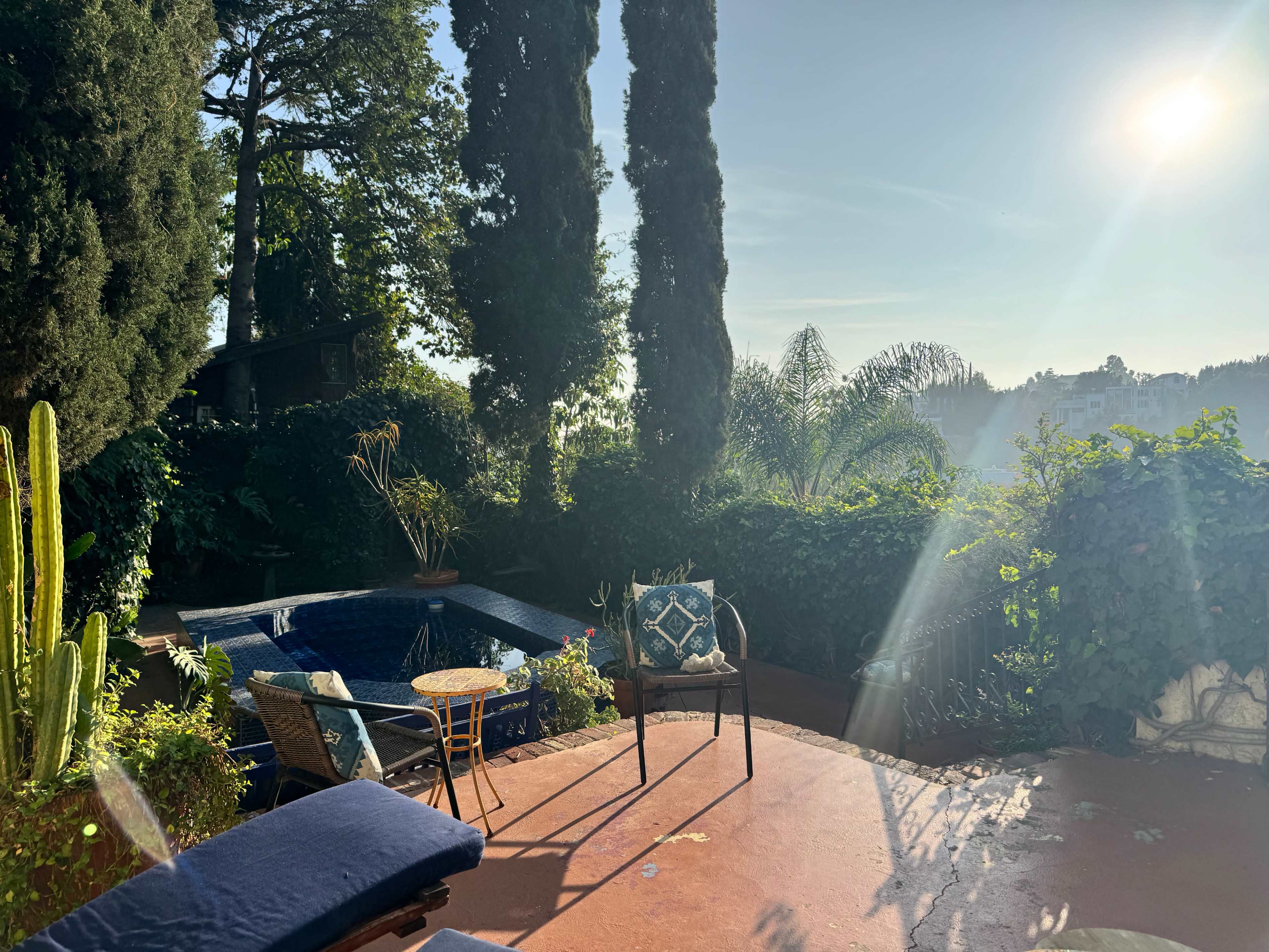 A sunlit patio features a pool surrounded by lush greenery and outdoor furniture.