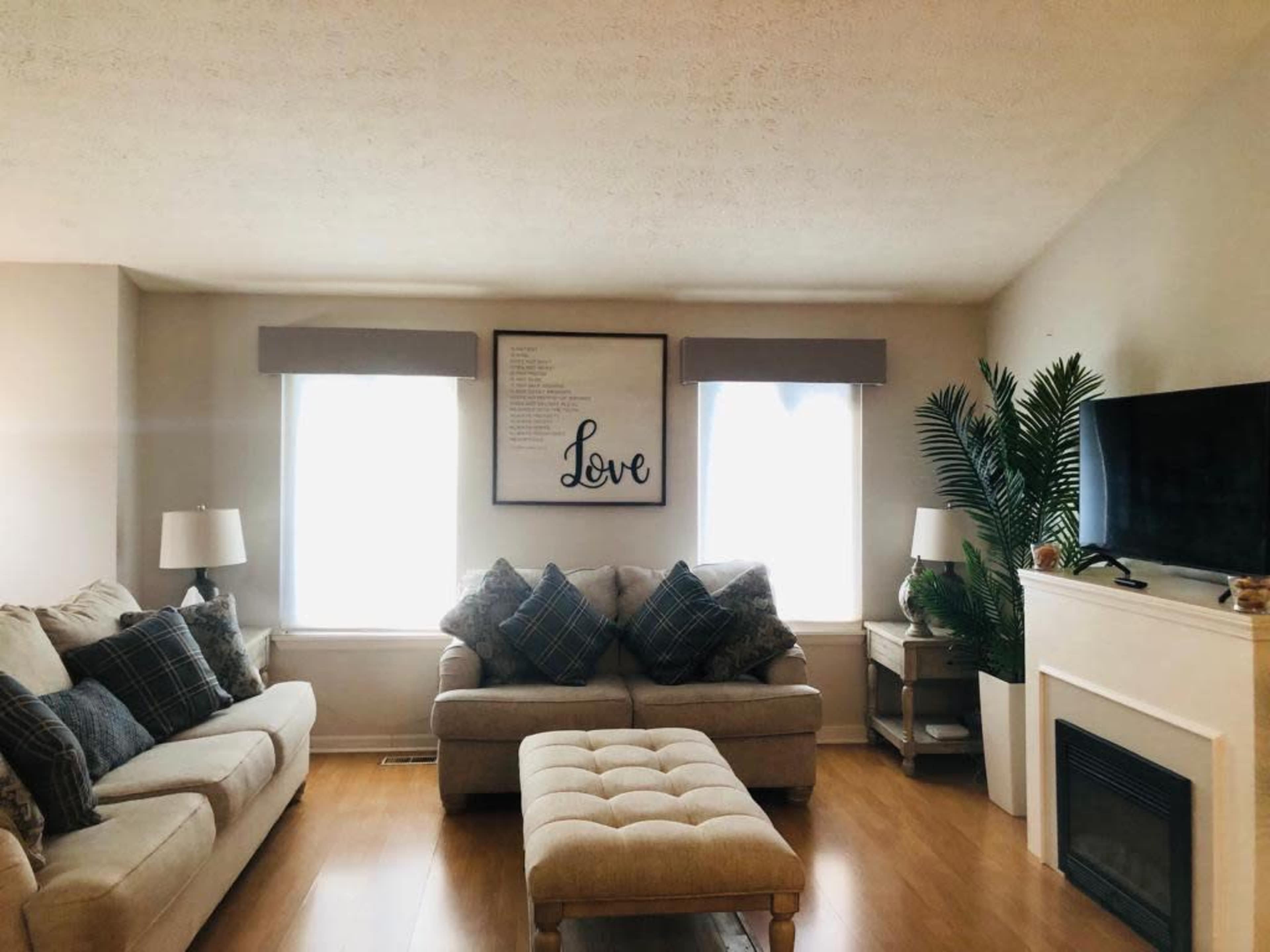 A living room features two windows with sheer coverings, a large framed "Love" sign on the wall, a light-colored sofa with patterned cushions, and a coffee table in front of a fireplace.