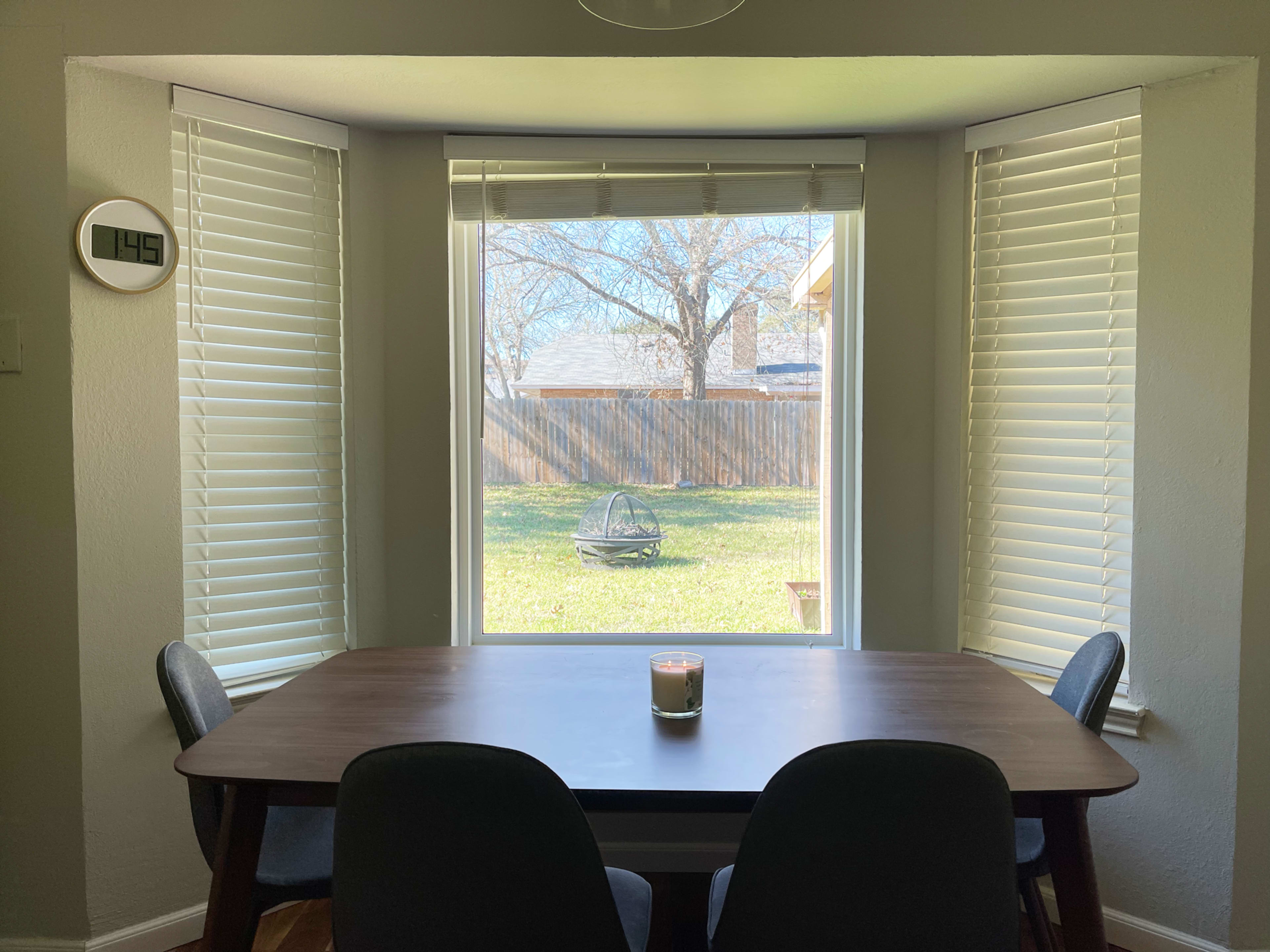 A table with a candle sits in a bright room, framed by large windows overlooking a backyard with a grassy area and a chair.