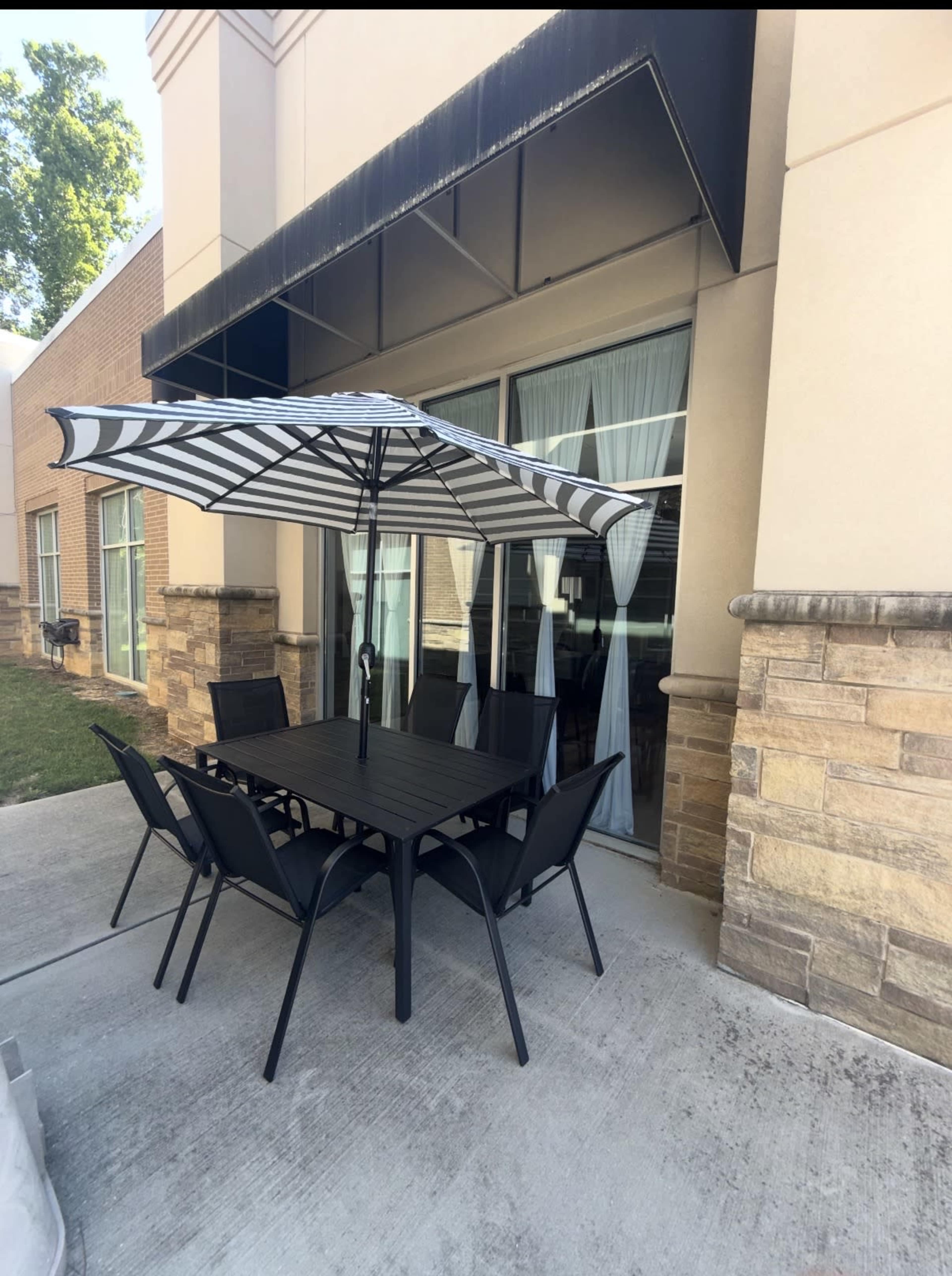 A black outdoor dining set with eight chairs and a striped umbrella is positioned on a concrete patio next to a building.