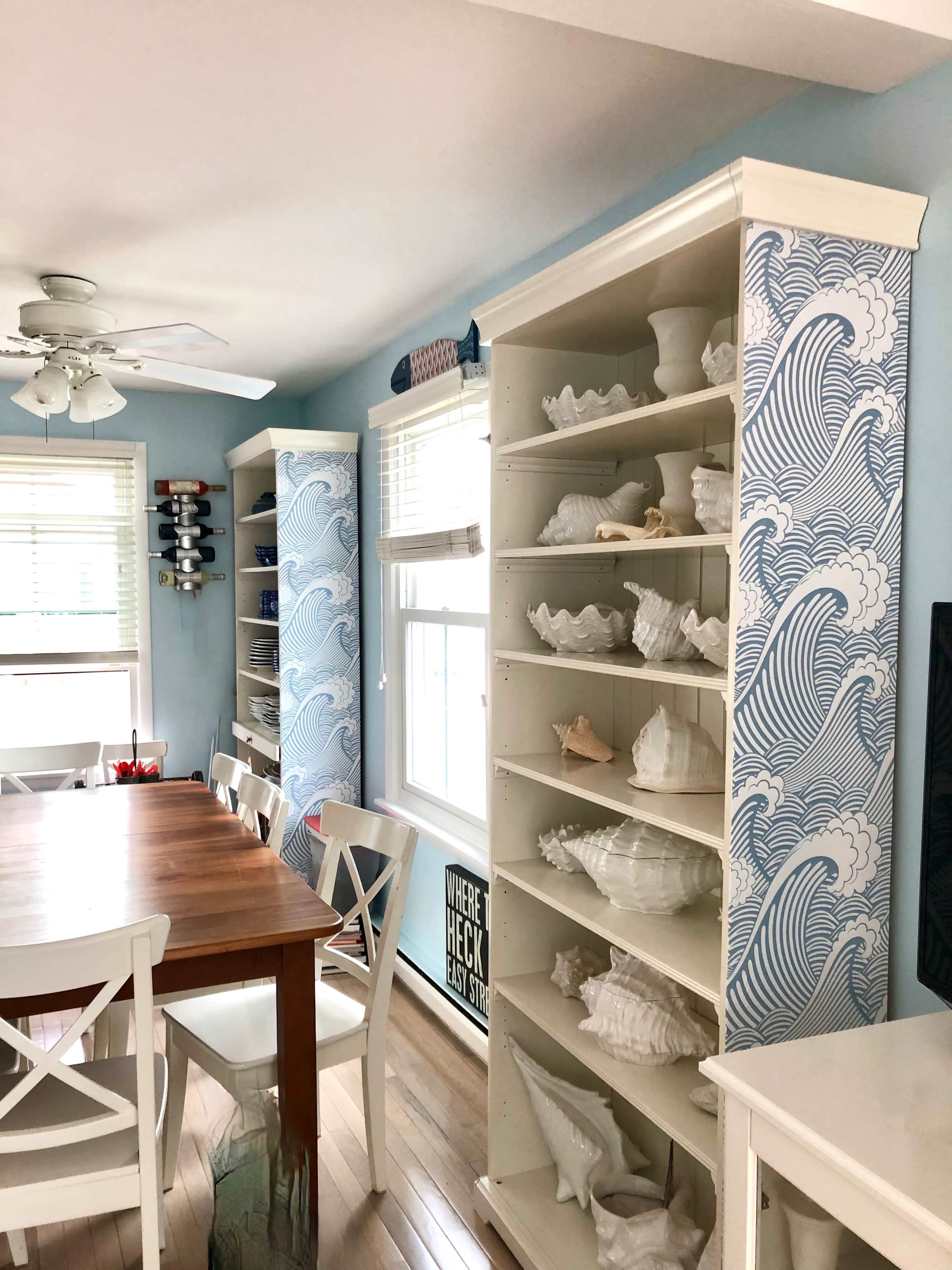 A dining area featuring a wooden table and two tall white bookshelves decorated with wave patterns and filled with seashell collectibles.