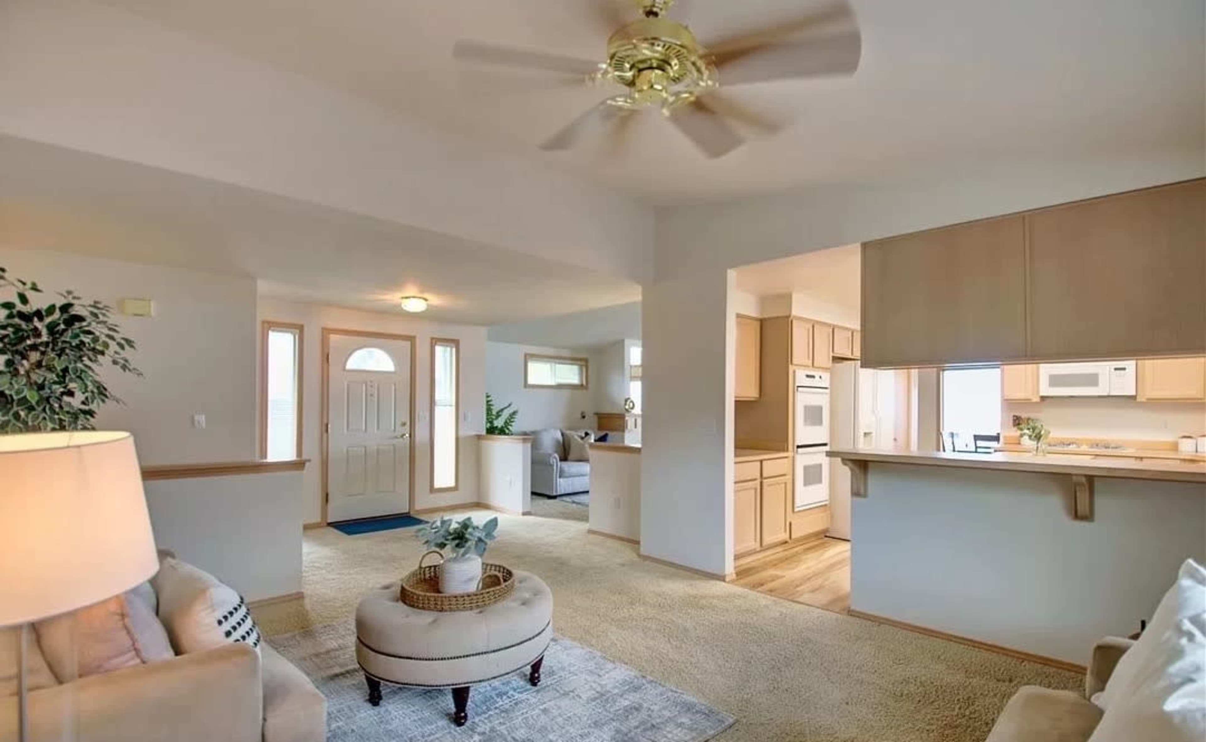 A well-lit living area with a ceiling fan, beige carpet, and an open layout connecting to the kitchen and entrance.
