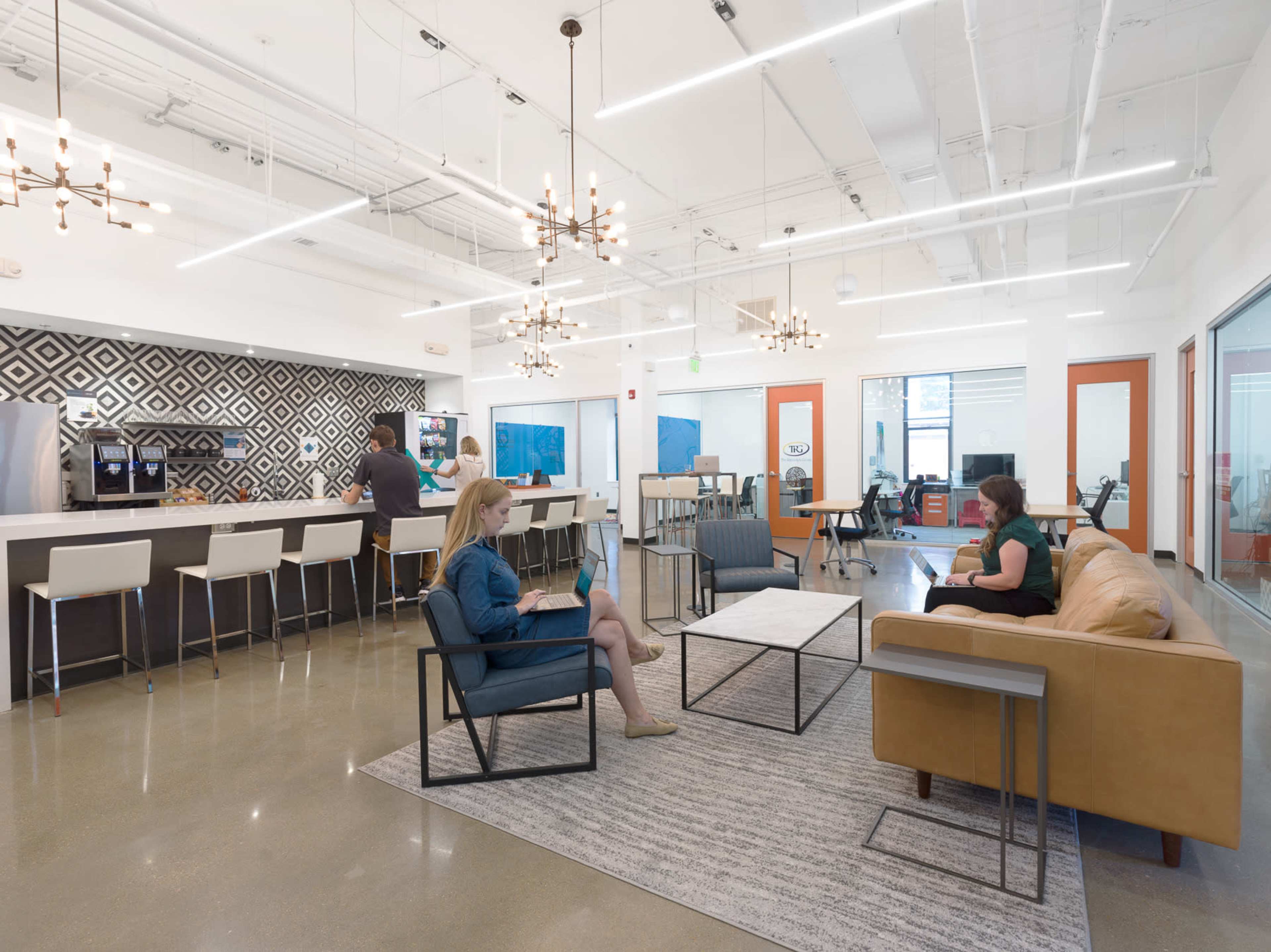 The image shows a modern office lounge area with a kitchen, featuring a bar, seating arrangements, and large windows.