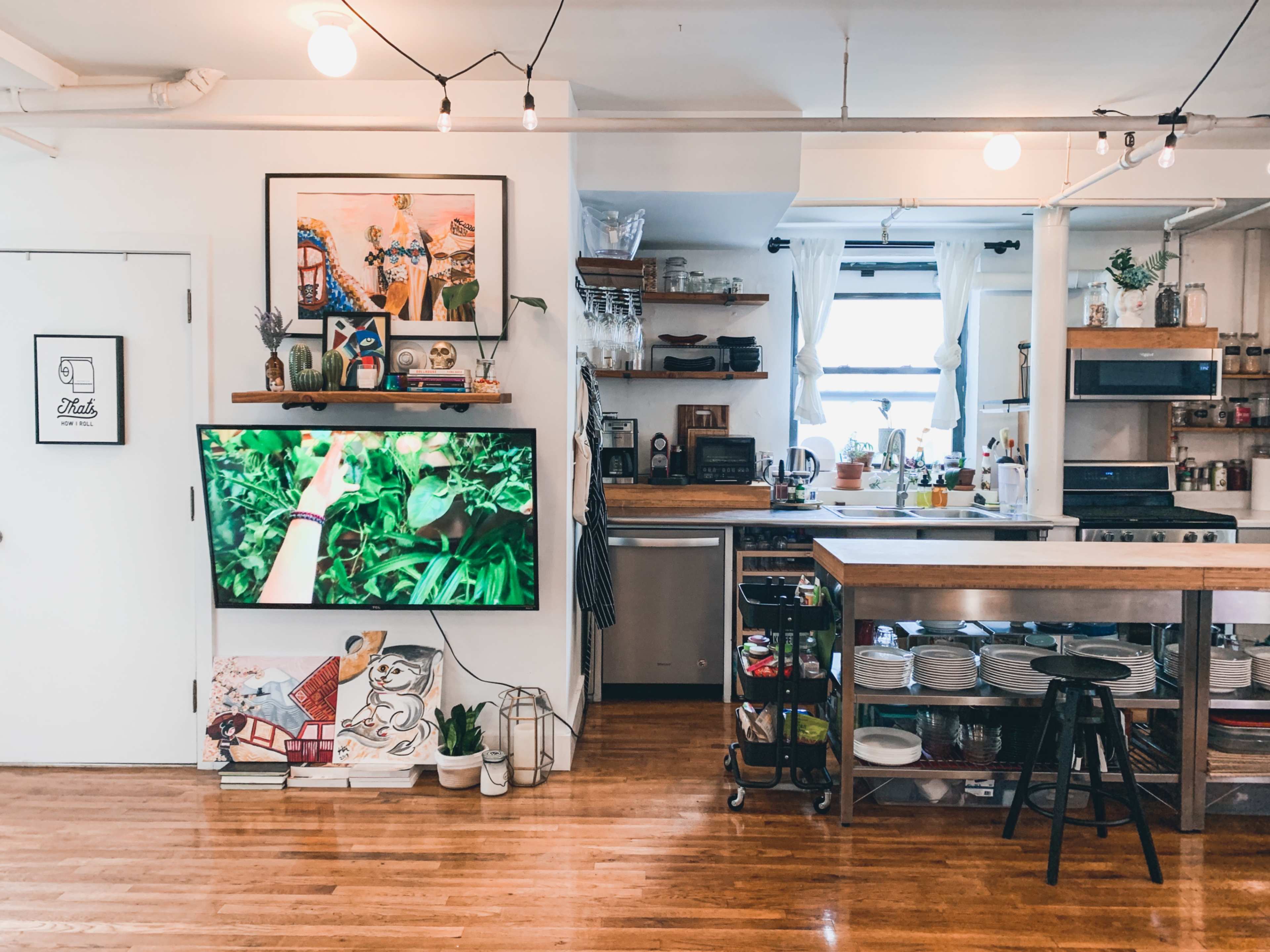 A modern kitchen and living area with a large television on one wall, a wooden kitchen counter, shelves of dishes, and various kitchen appliances in a well-lit space.