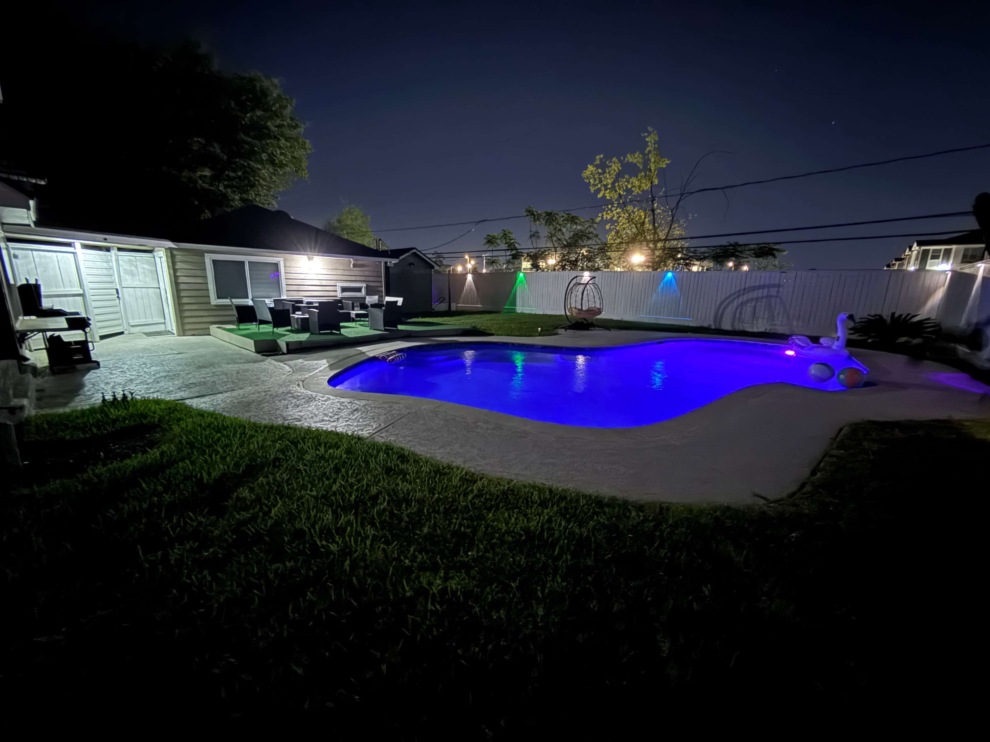 A backyard pool is illuminated with purple lighting at night, surrounded by grass and lounge furniture.