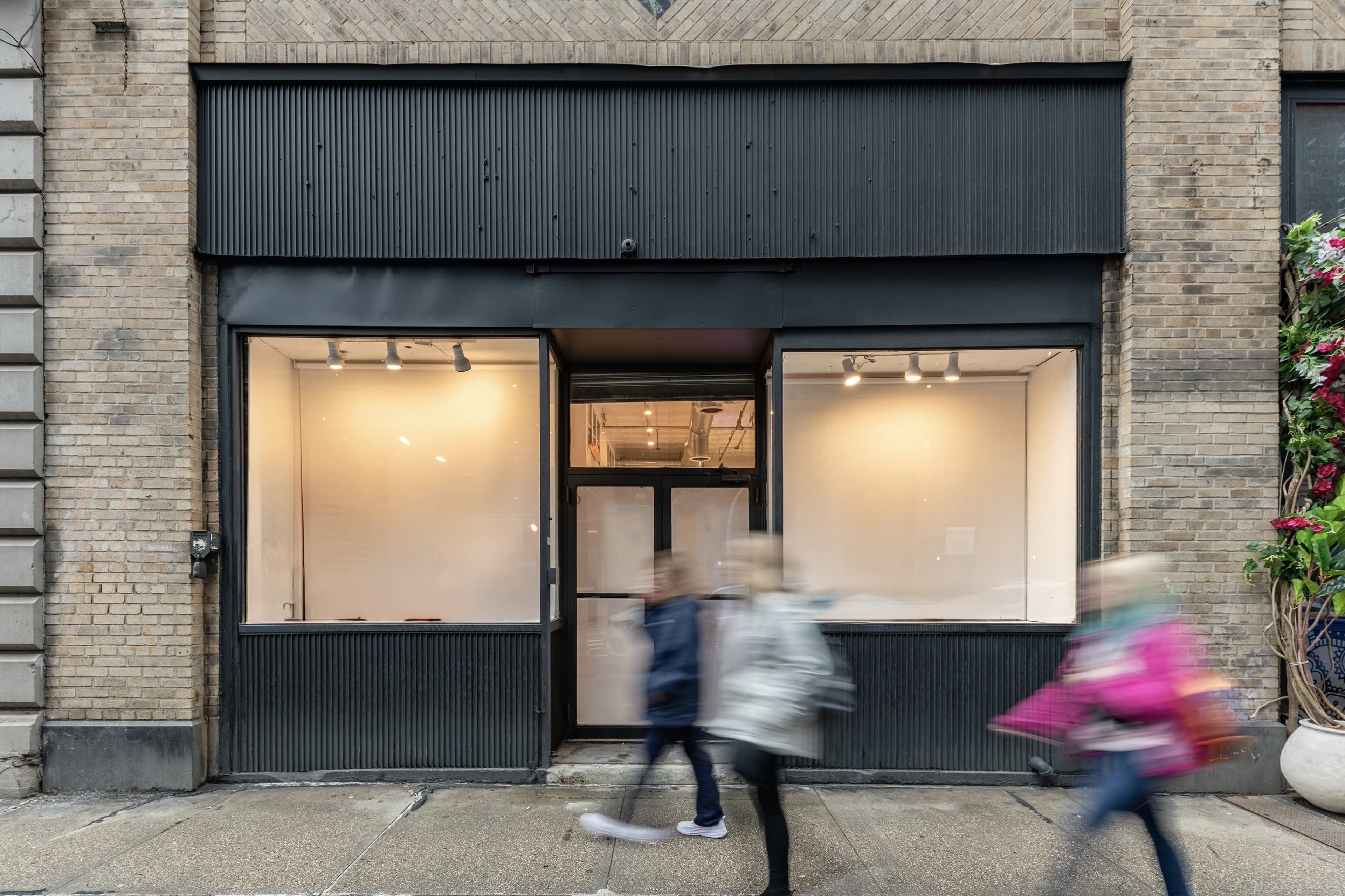 A storefront with large windows displays an open interior, while two people walk past on the sidewalk.
