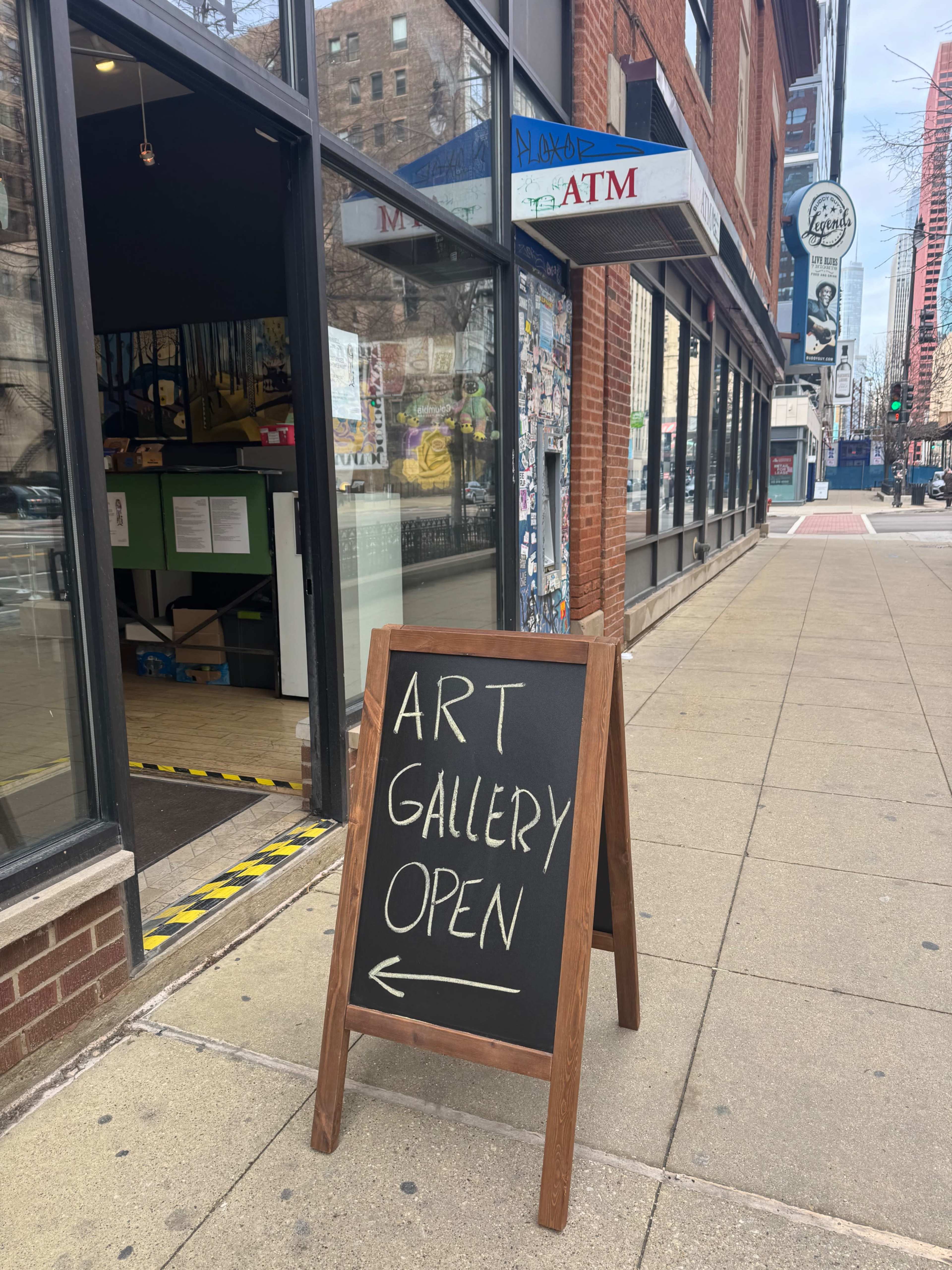 An A-frame sign displaying "ART GALLERY OPEN" is placed on the sidewalk outside an art gallery in an urban setting.