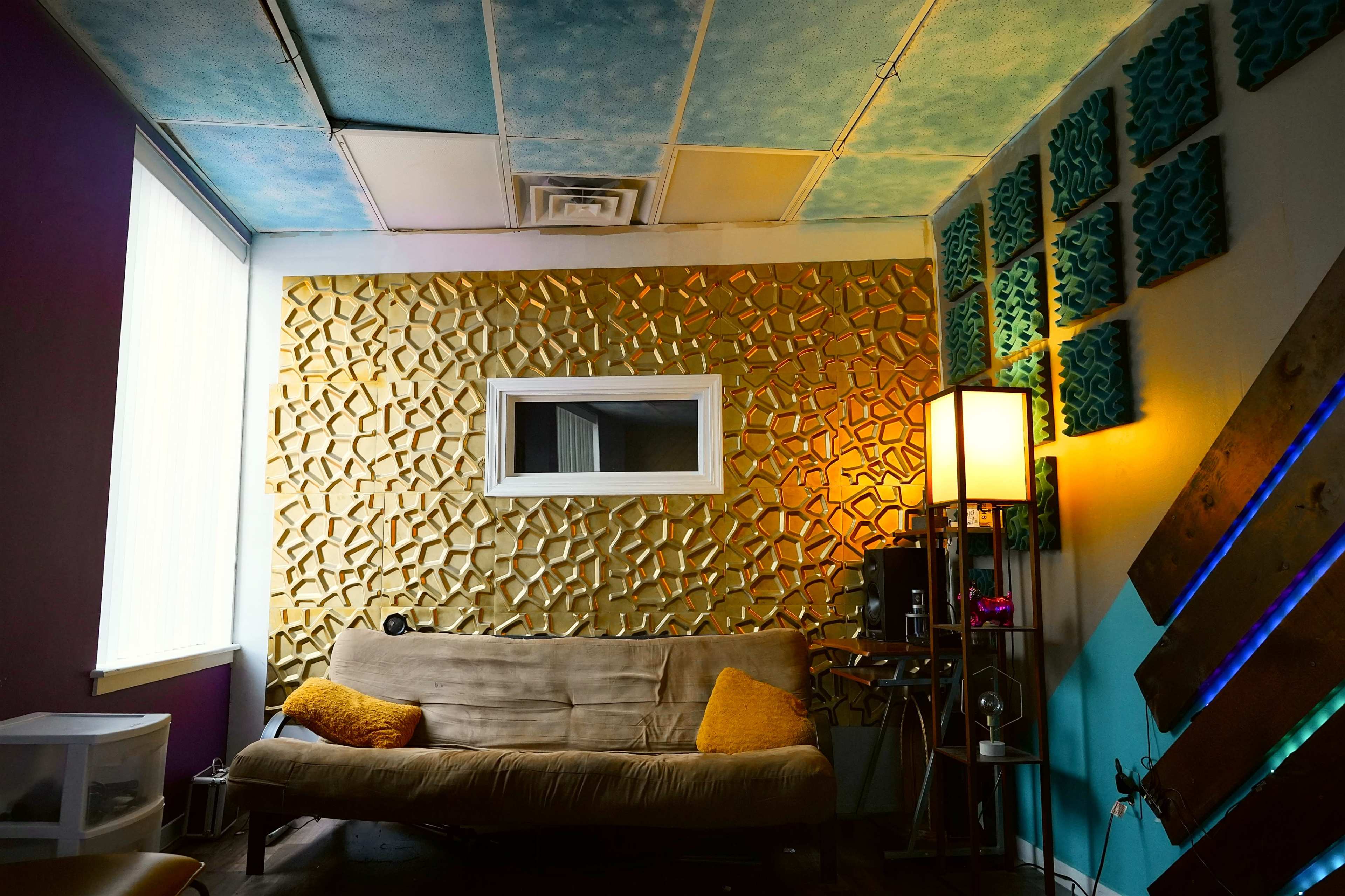 The image displays a small room featuring a textured gold accent wall, a beige couch with an orange cushion, and colorful acoustic paneling on the side walls.